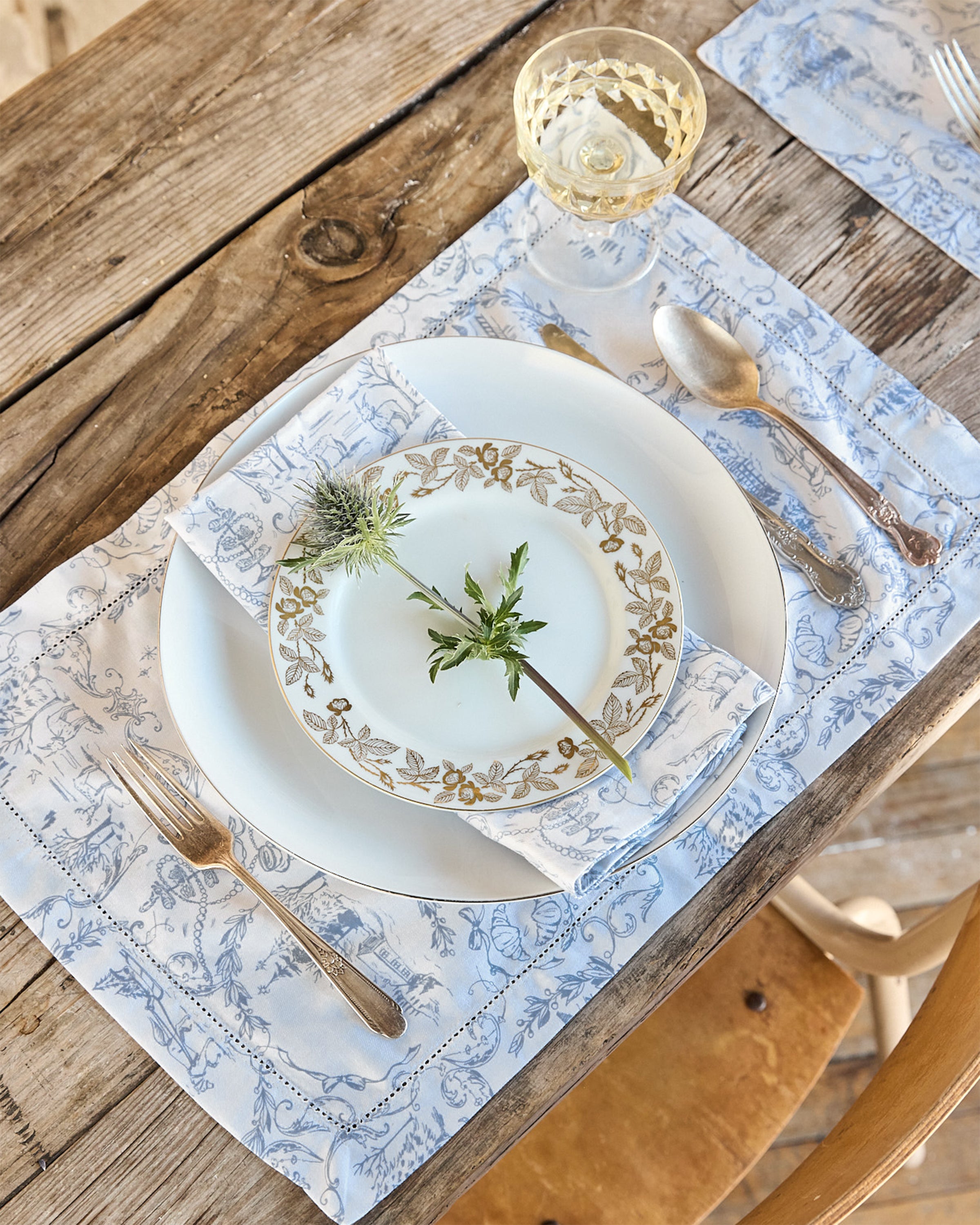 An elegant table setting on a rustic wooden table includes gold-trimmed plates with floral designs, sprigs of greenery, and 100% cotton linens. A crystal glass with a light drink sits atop the Petite Plumes limited-edition Maman x Petite Plume Alpine Toile Placemat to complete the look.