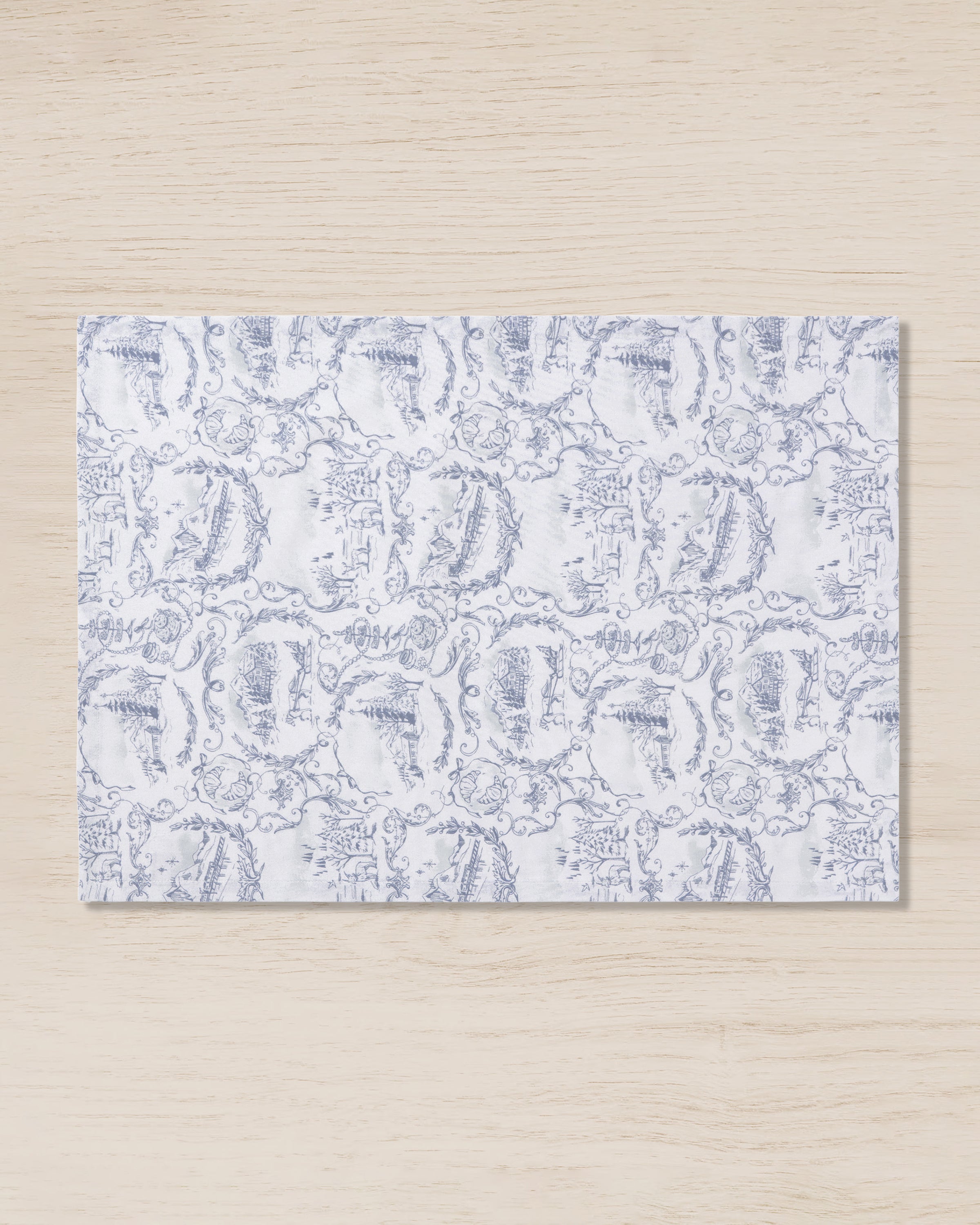 A light wood surface is adorned with a rectangular Maman x Petite Plume Alpine Toile Placemat by Petite Plume, featuring intricate limited-edition slate blue illustrations of diverse landscapes and buildings in a repeating pattern.