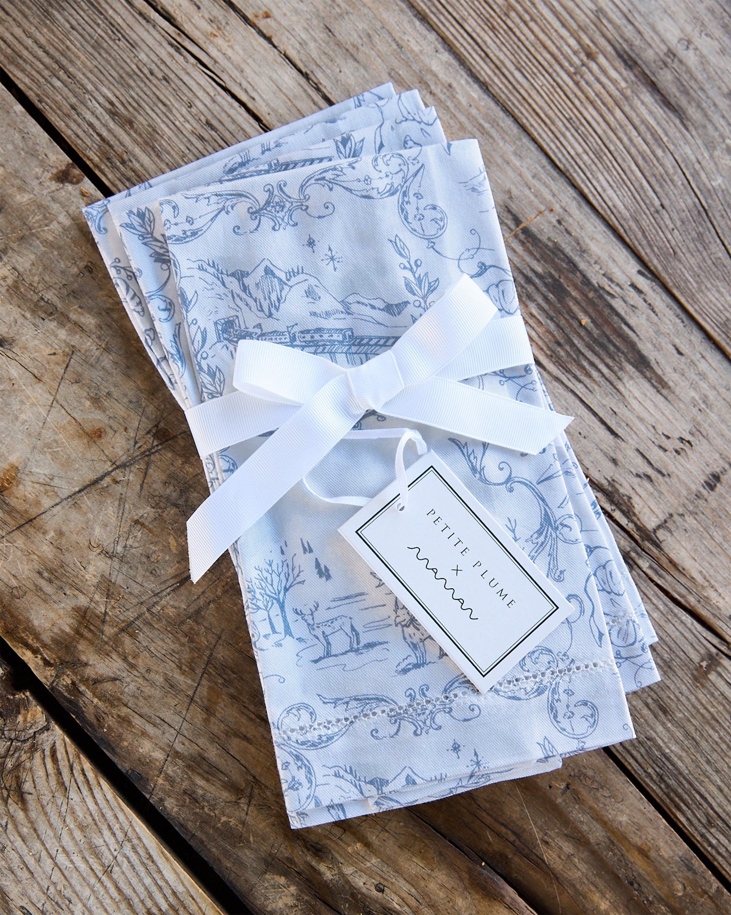 A limited-edition stack of Maman x Petite Plume Alpine Toile slate blue napkins, tied with a white ribbon and tag, rests on a rustic wooden table. Made from 100% cotton, these linens showcase delicate blue scenic designs ideal for any festive tablescape.