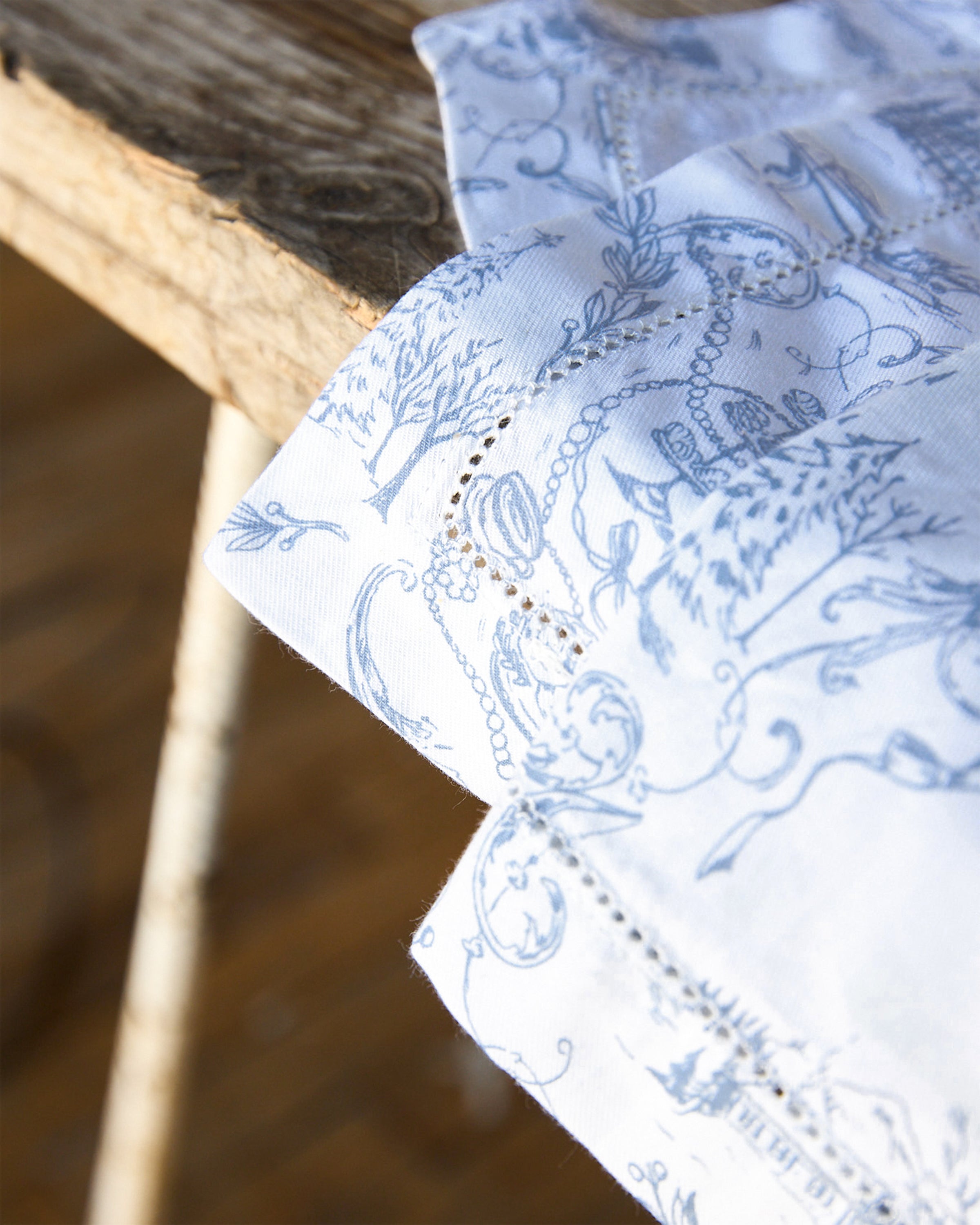 A Maman x Petite Plume Alpine Toile Placemat by Petite Plume, crafted from 100% cotton, features intricate blue floral and botanical patterns. Draped over an aged wooden chair, it contrasts beautifully to enhance any festive tablescape with elegance.