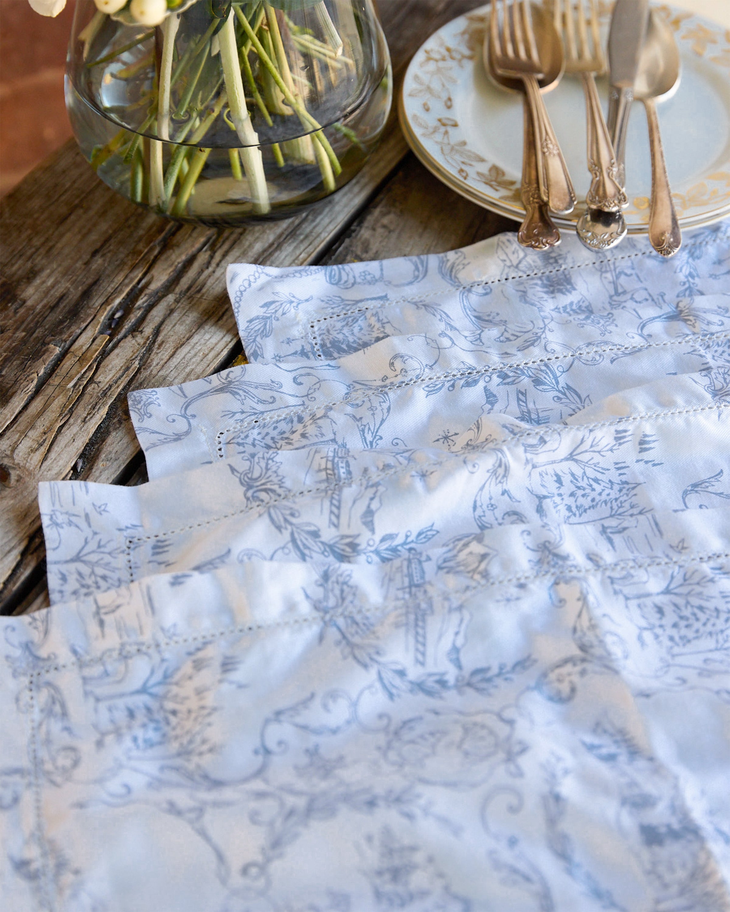 A rustic wooden table is adorned with Petite Plumes exclusive Maman x Petite Plume Alpine Toile Placemats, made of 100% cotton in limited-edition slate blue, paired with ornate plates, silver cutlery, and a glass vase containing white flowers on green stems.