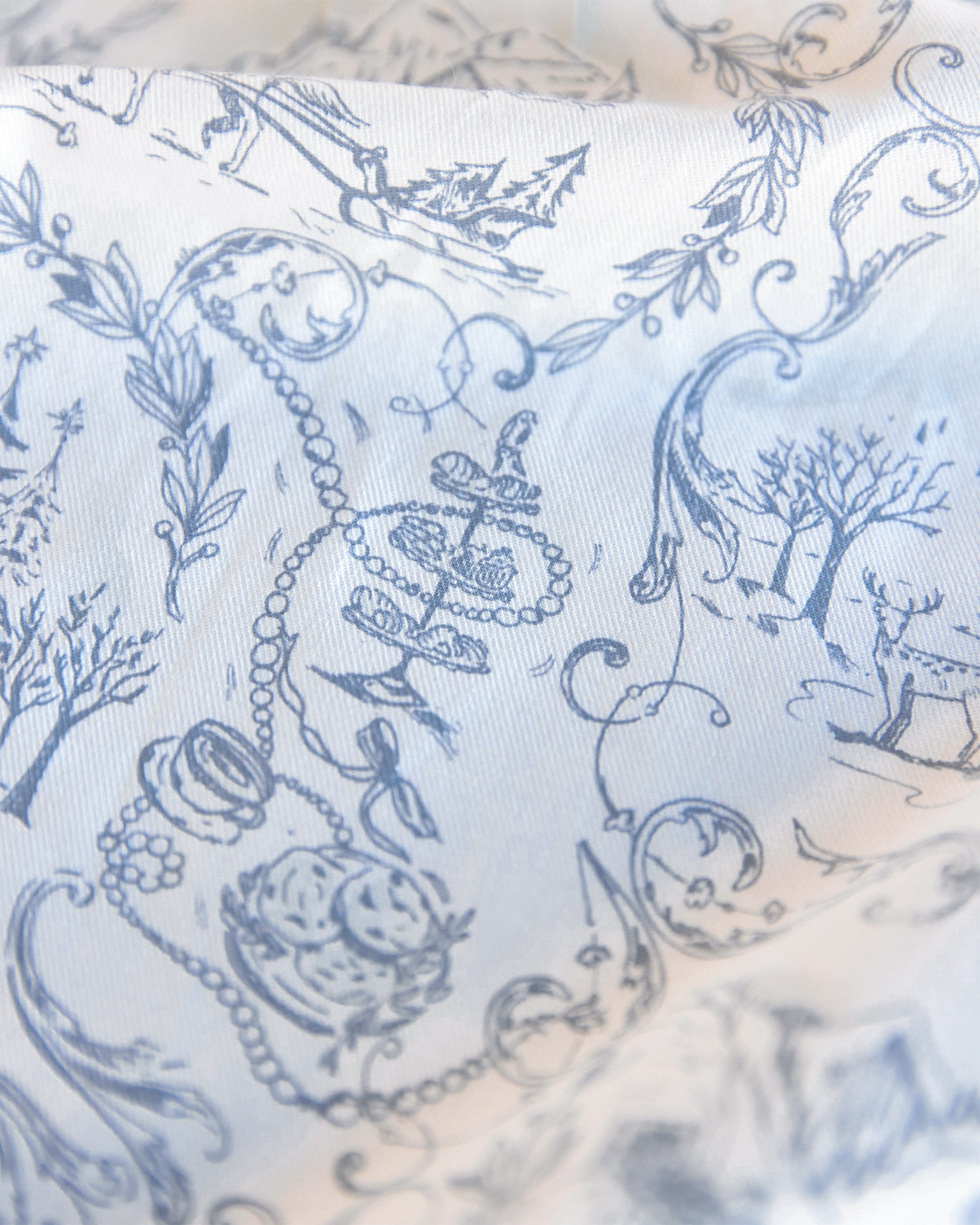 A close-up of the Maman x Petite Plume Alpine Toile Placemats by Petite Plume showcases an intricate slate blue toile design on 100% cotton, featuring trees, a boat with an anchor, pearls, and decorative swirls. These limited-edition linens offer a vintage elegance ideal for festive tablescapes.