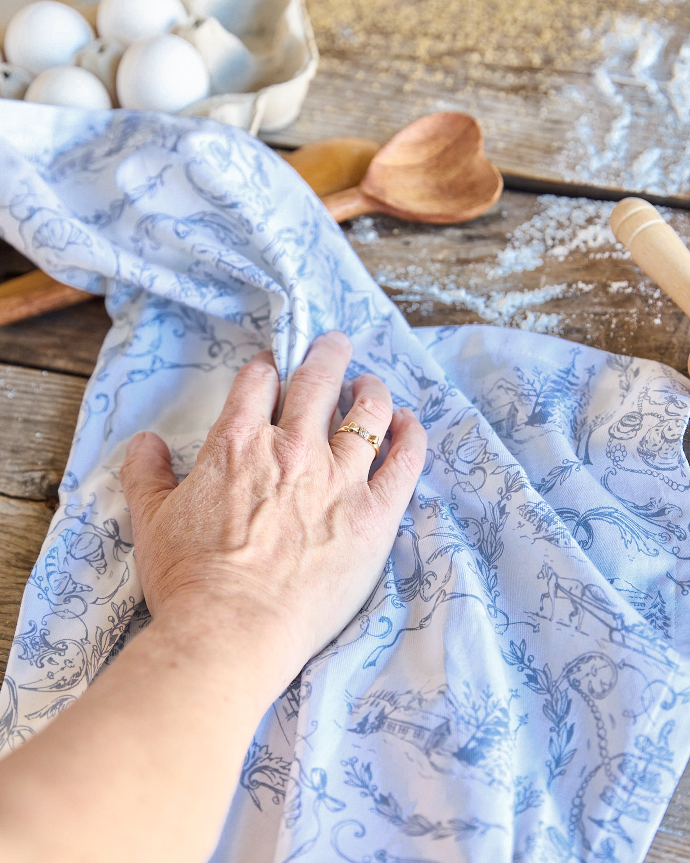 A hand adorned with a ring rests on Maman x Petite Plume Alpine Toile Tea Towels. Surrounding it are eggs in a carton, a wooden spoon, a rolling pin, and spilled flour on a wooden surface.