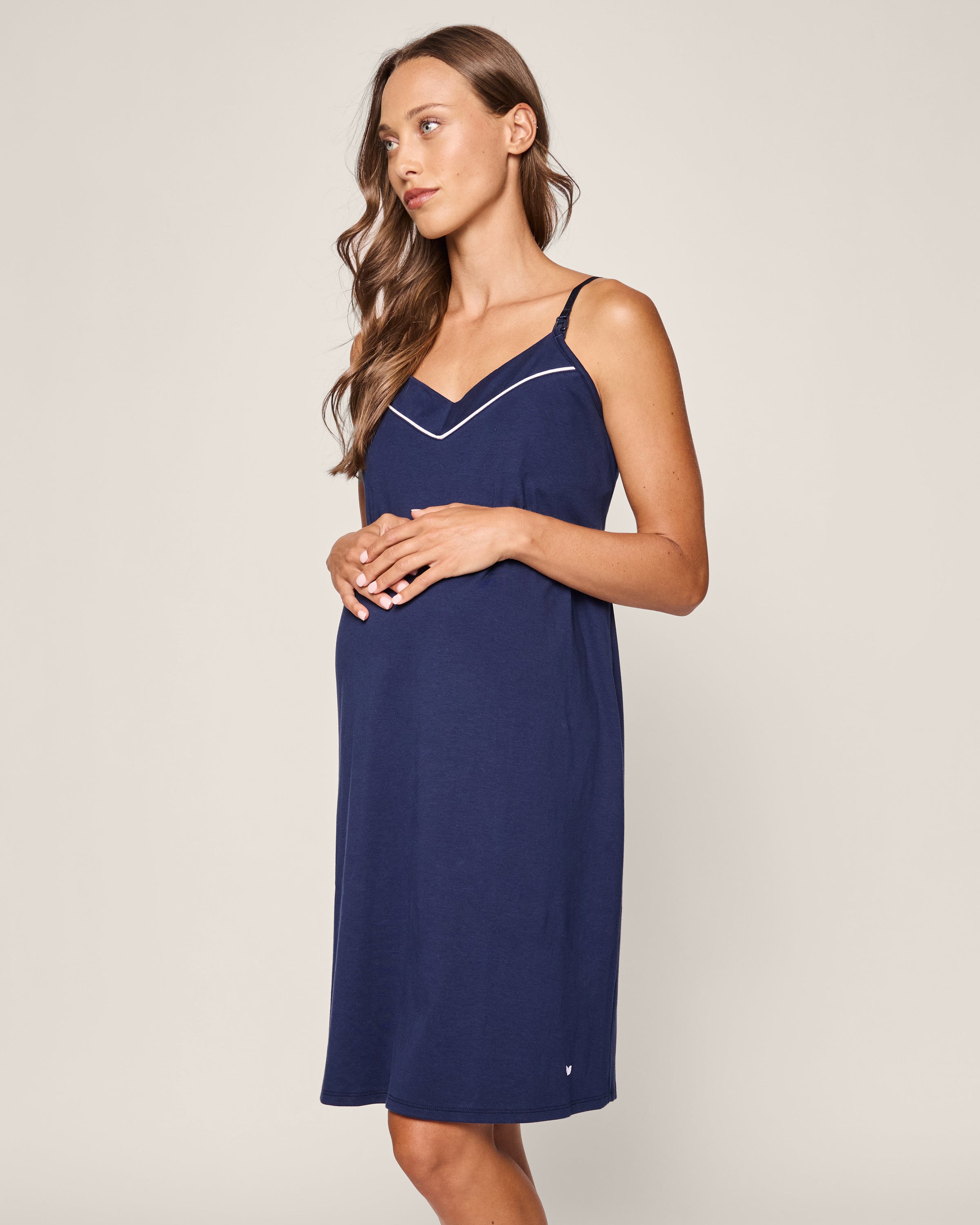 Against a neutral backdrop, a woman with cascading long wavy hair cradles her belly, wearing Petite Plumes Womens Pima Maternity Nightgown in Navy.