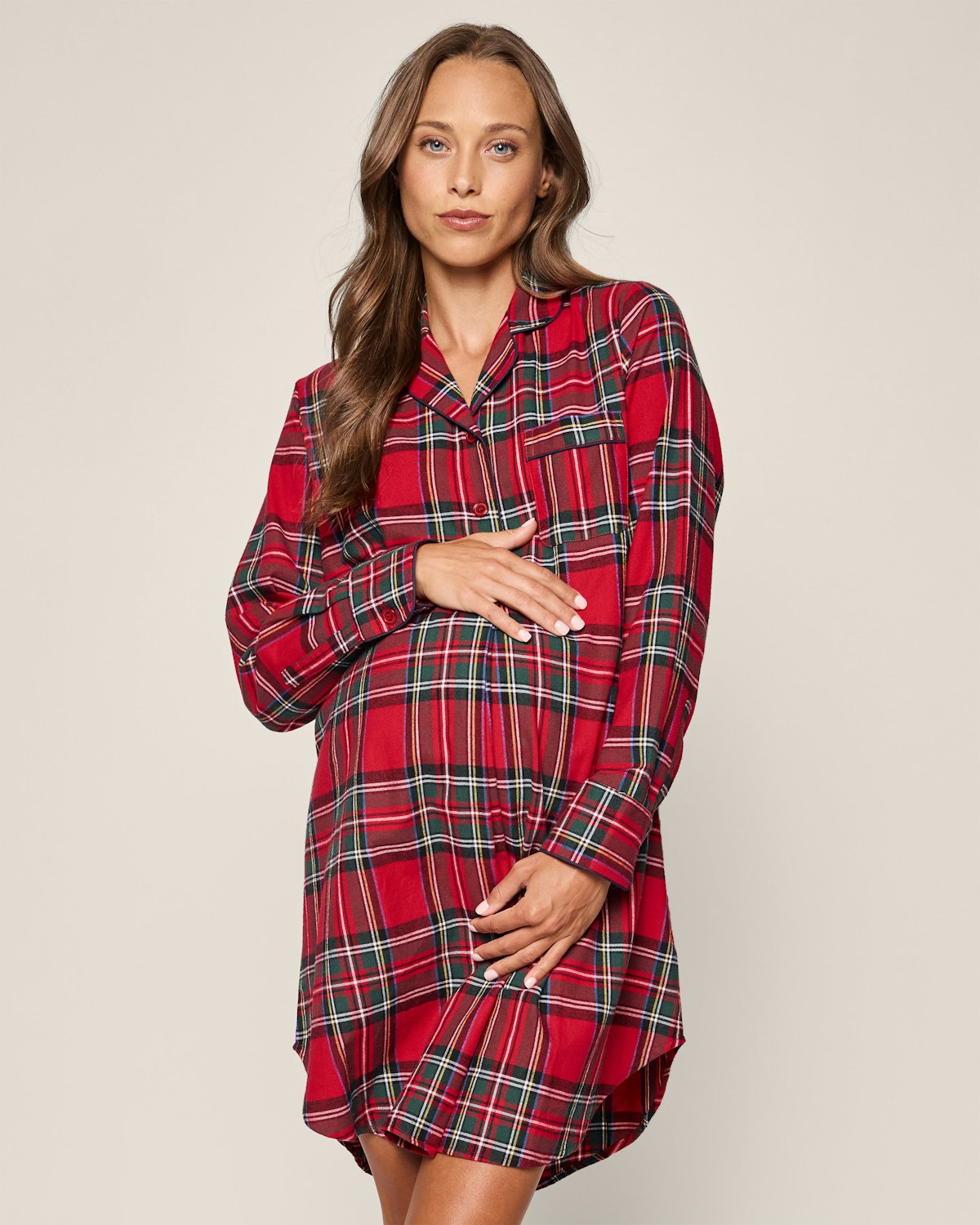 Against a plain backdrop, a woman with long hair gently rests her hand over her stomach while wearing Petite Plumes Womens Brushed Cotton Maternity Nightshirt in Imperial Tartan and gazing forward calmly.