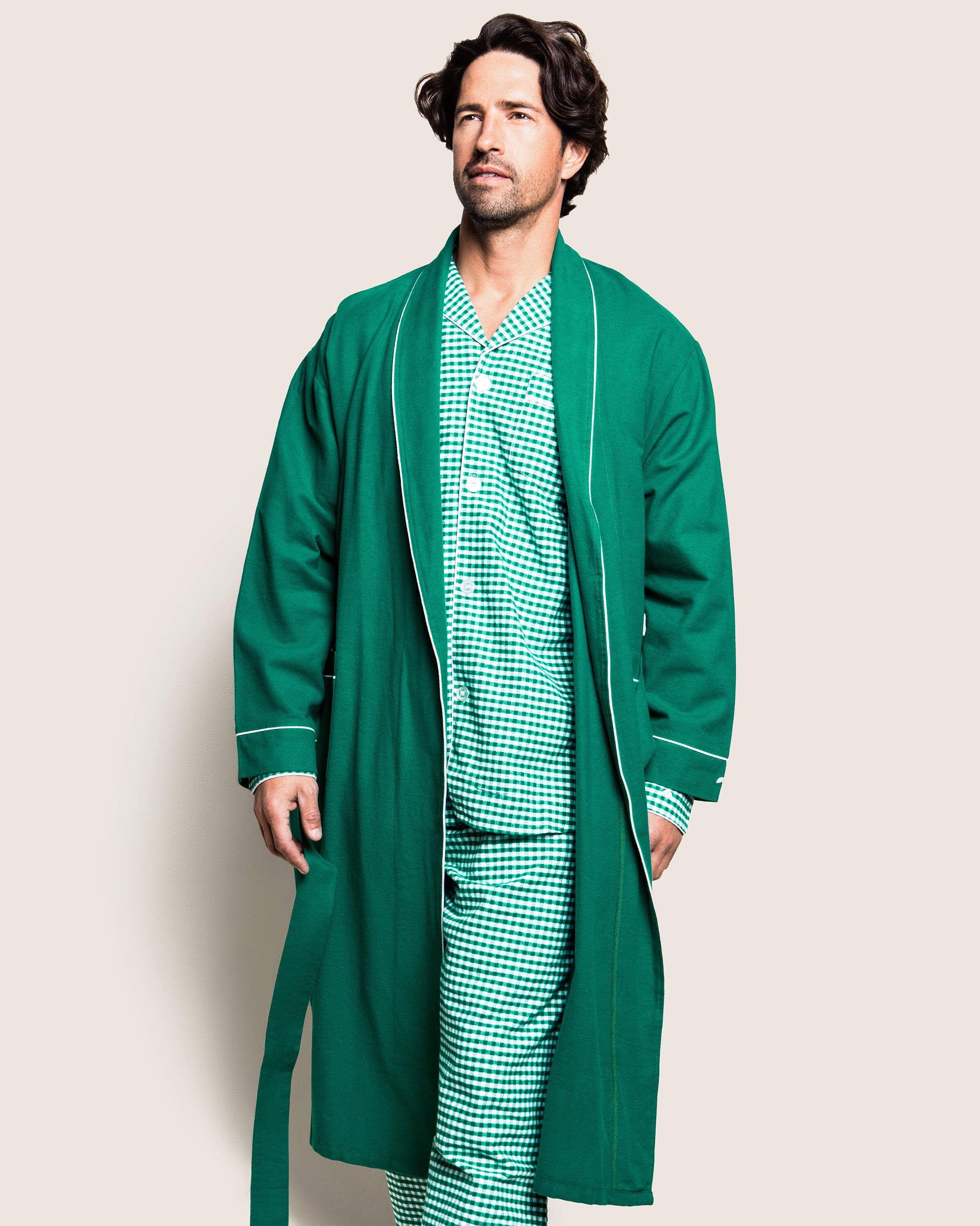 A man confidently wears a Petite Plume Mens Flannel Robe in Forest Green with white piping over a checkered pajama set. This luxurious 100% cotton robe complements the comfortable outfit against a plain light beige background.