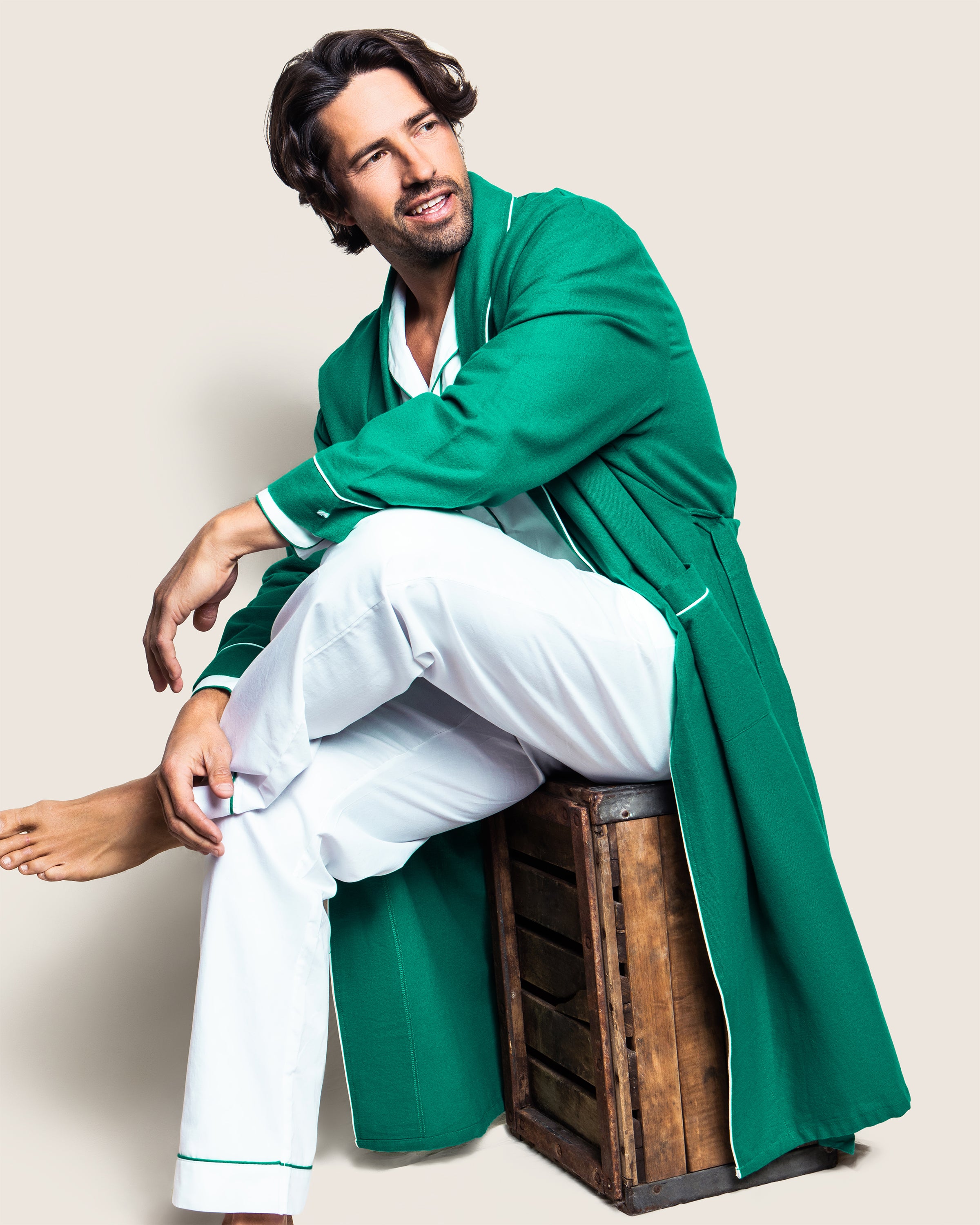 A man sits on a wooden crate, wrapped in Petite Plumes Mens Flannel Robe in Forest Green with White Piping over white clothing, looking thoughtful against a beige background.