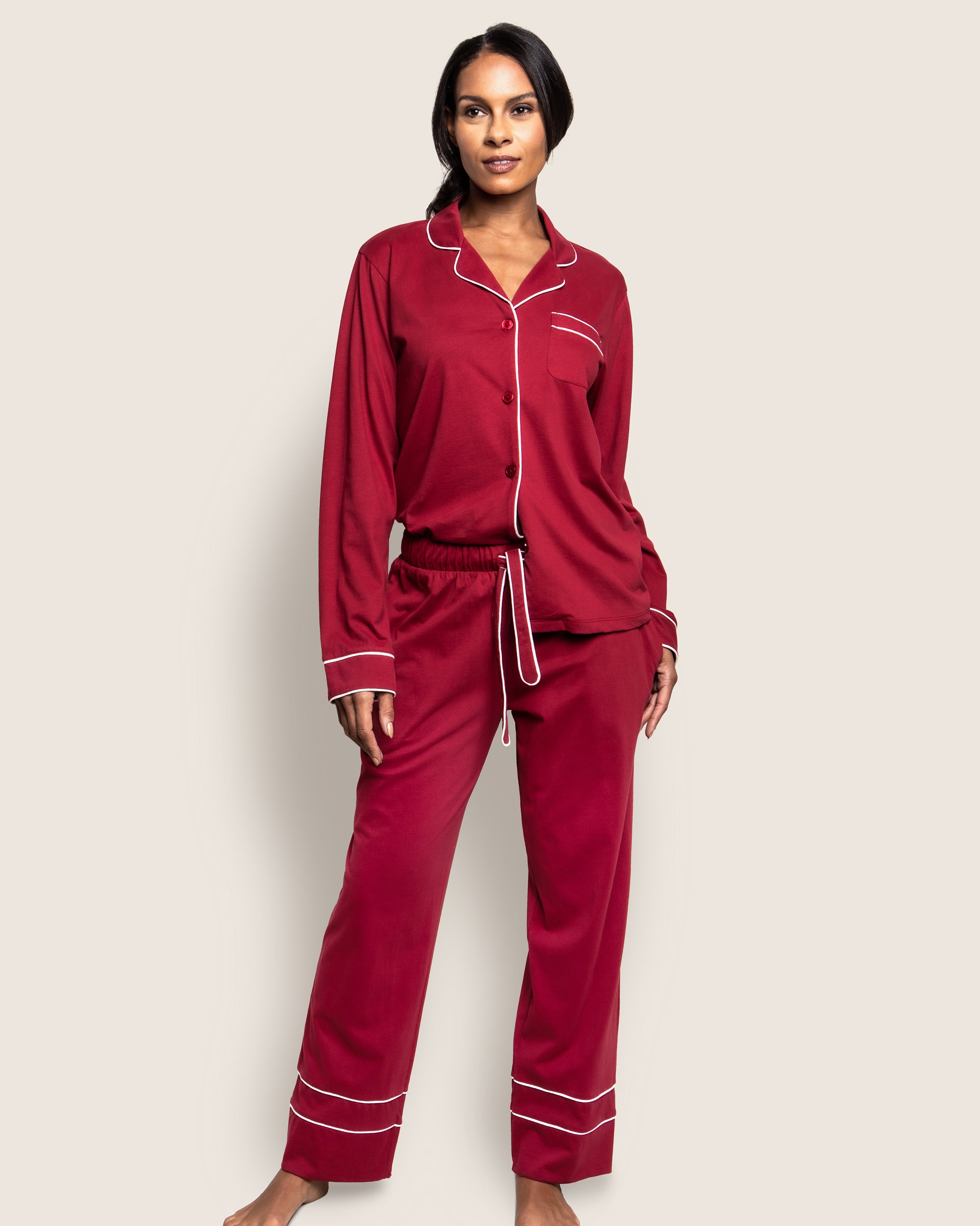 A person wears the Womens Pima Astaire Pajama Set in Bordeaux by Petite Plume. This luxurious sleepwear, made from buttery Peruvian Pima cotton, includes a button-up top with white piping and elastic-waist pants. The light background highlights the cozy elegance of this breathable ensemble.