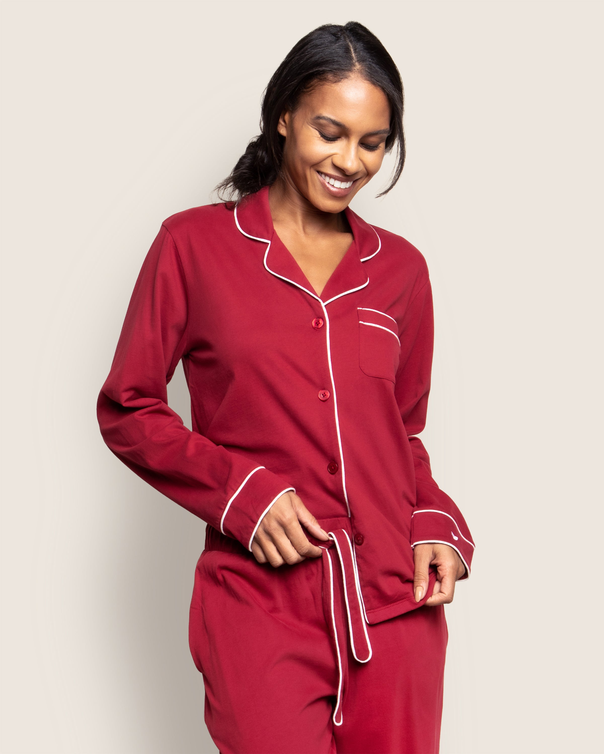 A person smiles while wearing Petite Plumes Womens Pima Astaire Pajama Set in Bordeaux, featuring white piping. The buttery breathable set includes a buttoned top with a chest pocket as they adjust the waistband of their pants against a light, solid background.