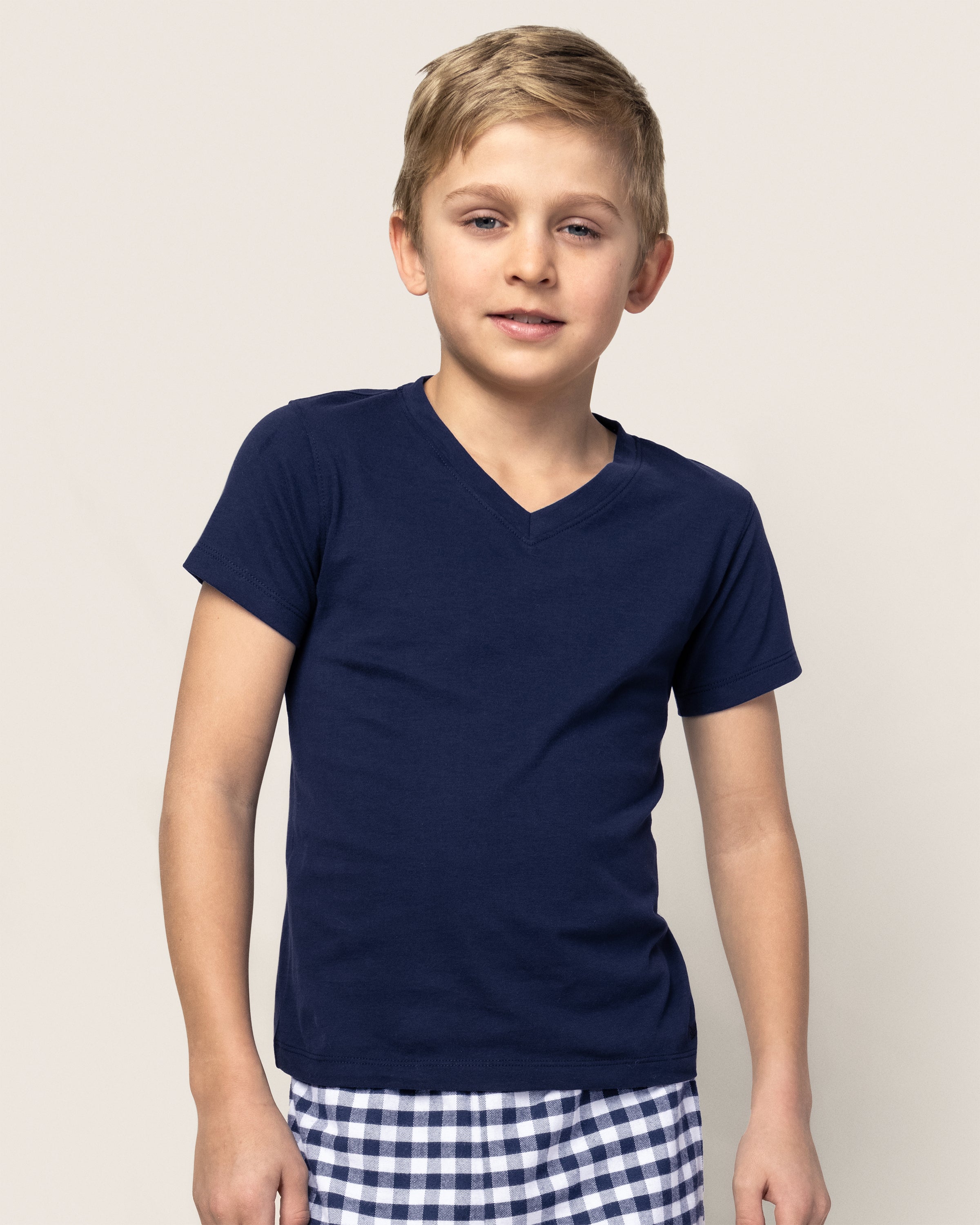 A young boy with short blond hair is wearing a Petite Plume Kids Pima V-Neck Top in Navy and checkered shorts. He stands against a plain light-colored background, looking towards the camera with a neutral expression.