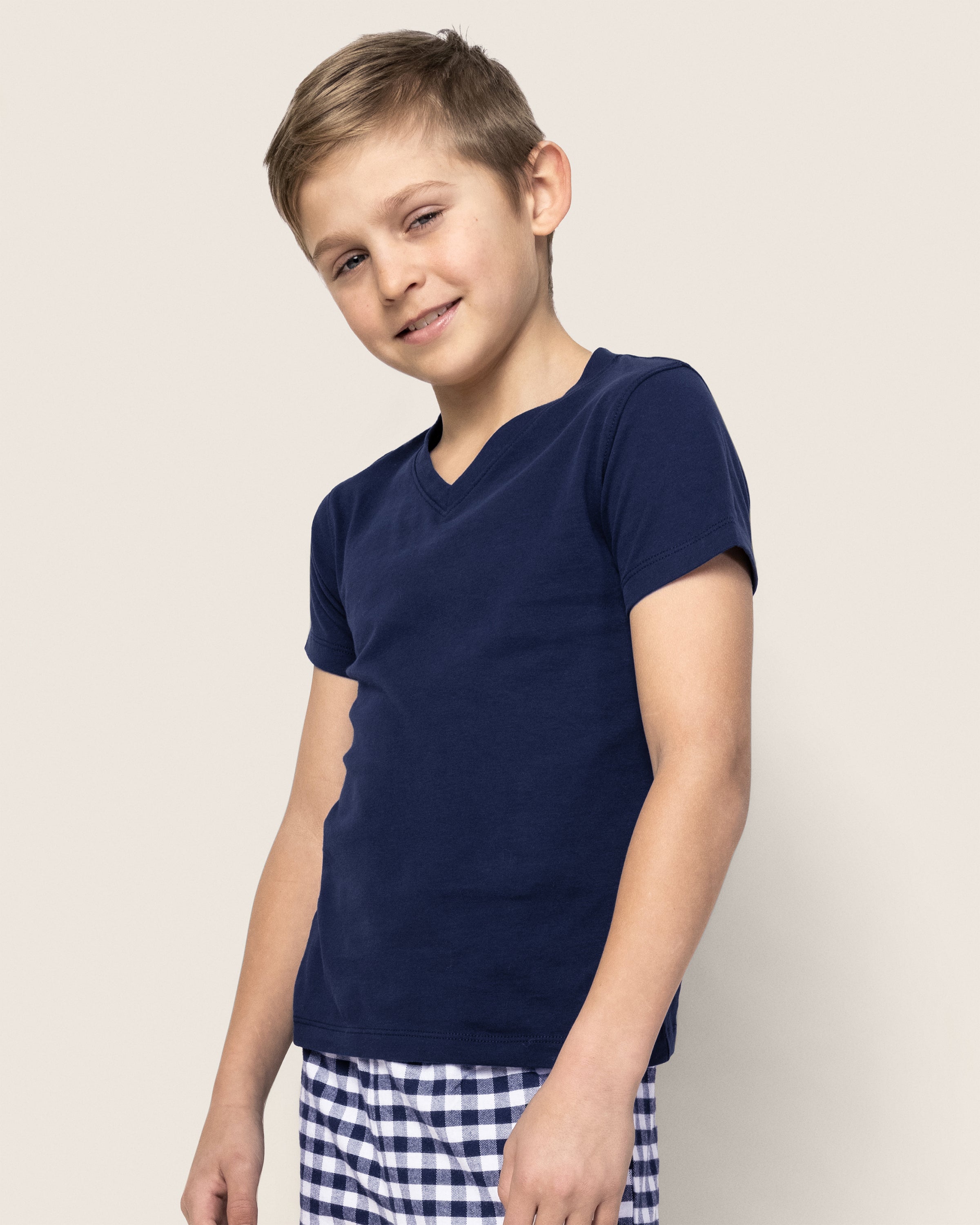 A young boy smiles wearing the Kids Pima V-Neck Top in Navy from Petite Plume, paired with checkered pants made of soft, yarn-dyed Peruvian Pima cotton. The neutral background highlights his casual attire and relaxed pose, evoking cozy jersey knit pajamas.