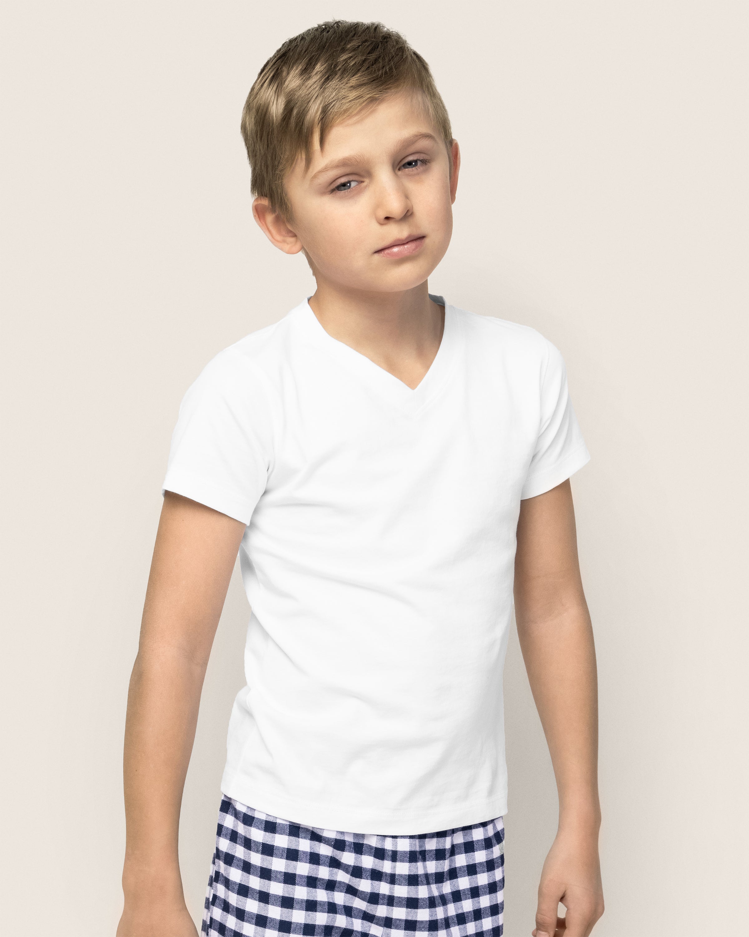 A young boy with short hair wears a Petite Plume Kids Pima V-Neck Top in white and blue checkered shorts. He stands against a light beige background, slightly leaning to one side with a calm expression.