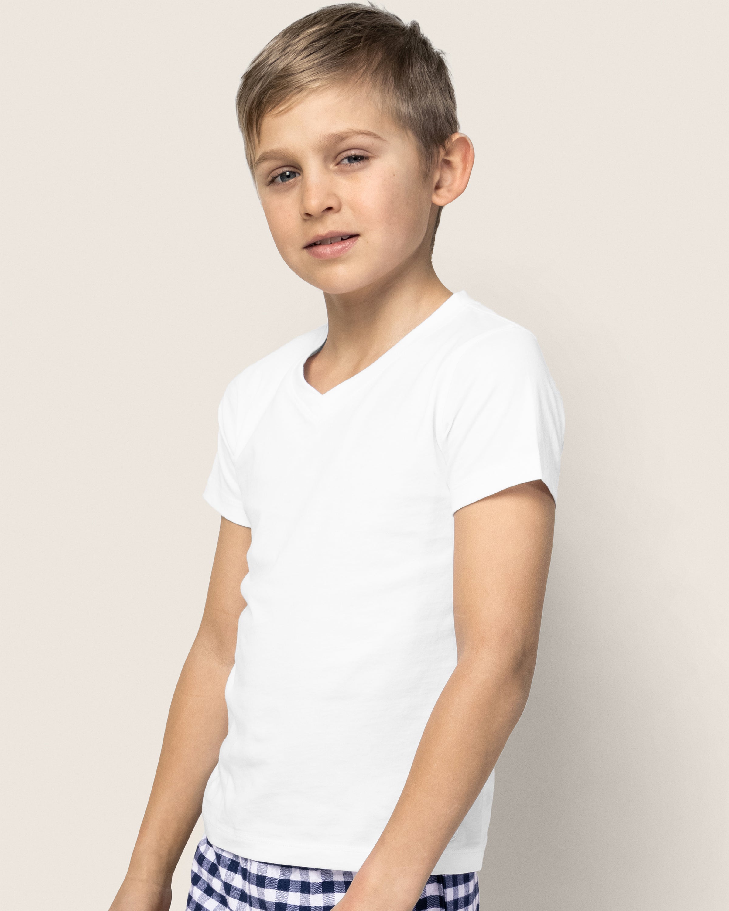 A young boy, wearing Petite Plumes Kids Pima V-Neck Top in White and blue checkered pants made of Peruvian Pima cotton, stands against a light background. His short, light brown hair complements his neutral expression, exuding timeless charm.