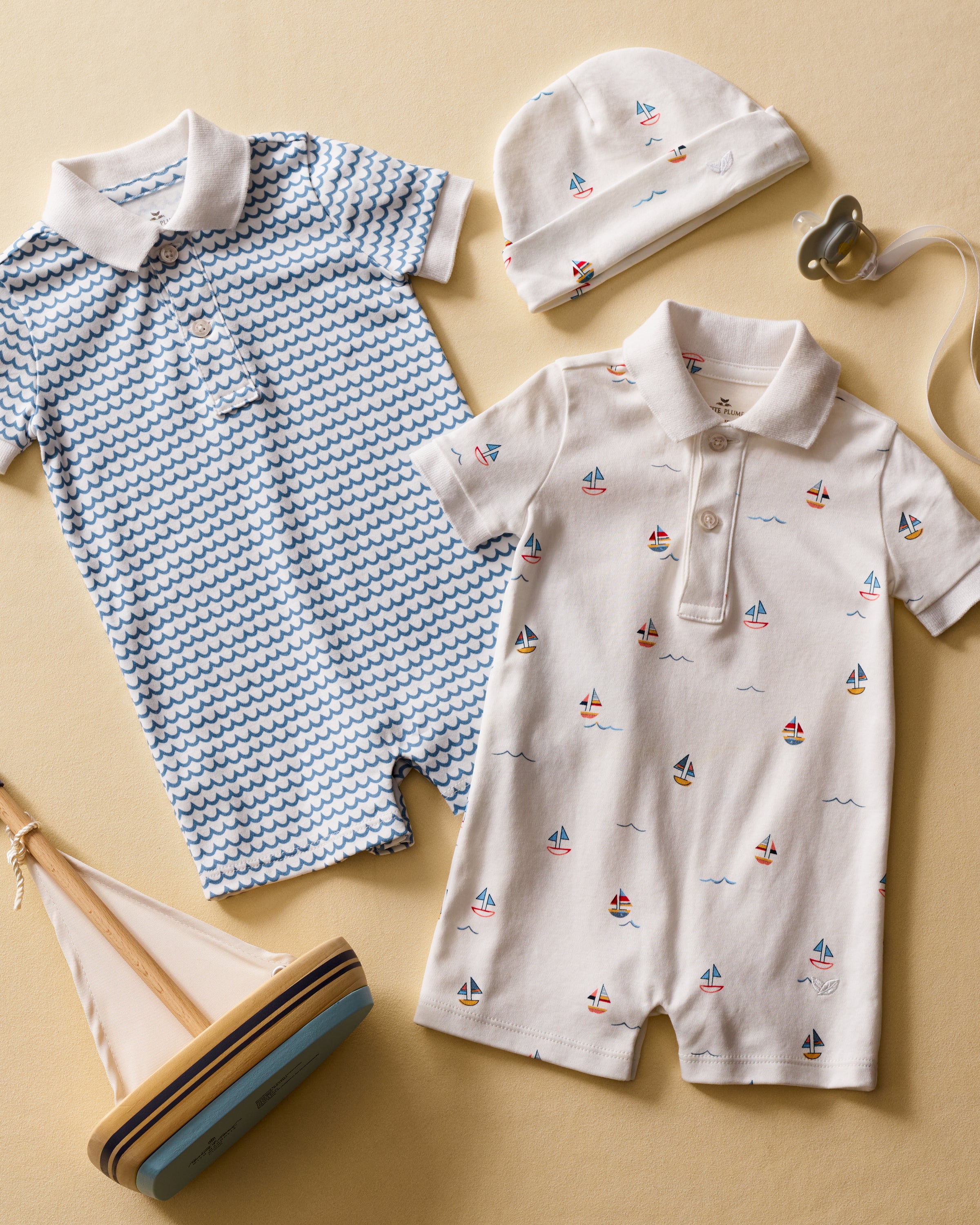 Laid out on a beige surface, the ensemble includes two collared rompers: a blue wave print and the Babys Pima Louis Play Romper in Bateau by Petite Plume, plus a matching sailboat-patterned hat, pacifier, and toy sailboat—all in charming 100% Pima cotton.