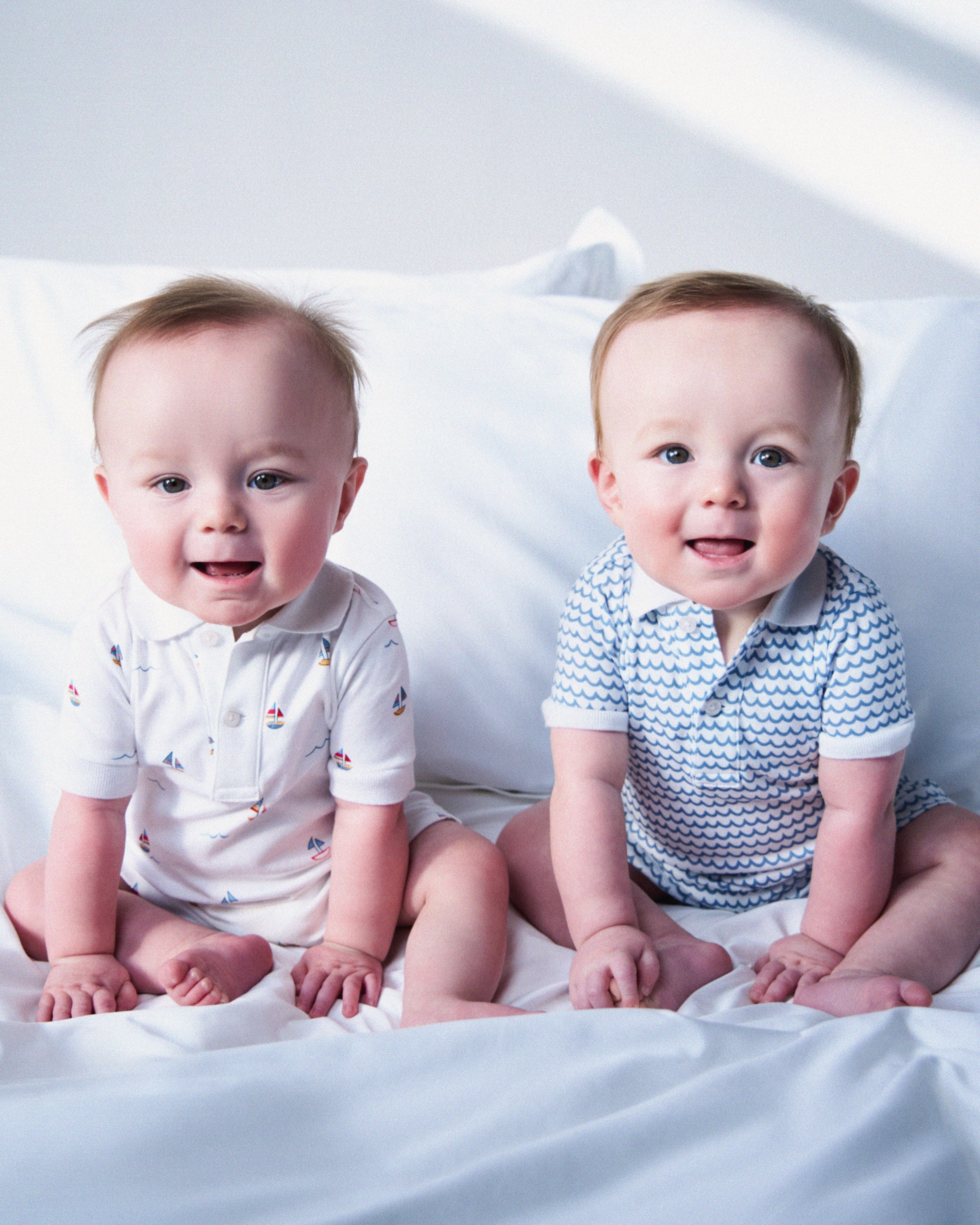 Two happy babies with light brown hair sit closely on a white bed, dressed in charming Petite Plume attire. One wears the Babys Pima Louis Play Romper in Bateau featuring a sailboat pattern, and the other sports a blue wave design, both smiling brightly in short-sleeved collared shirts.