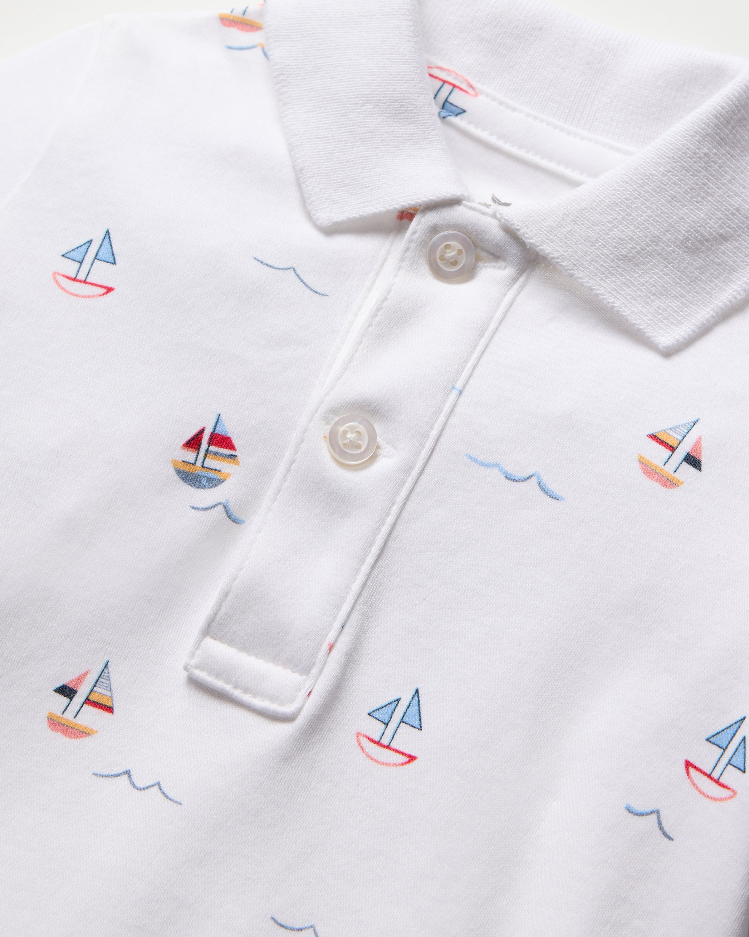 The Babys Pima Louis Play Romper in Bateau by Petite Plume is crafted from 100% Pima cotton, featuring a playful print with sailboats in blue, red, and yellow. It includes a classic collar and two-button placket, highlighted by wavy blue lines among the sailboat designs.