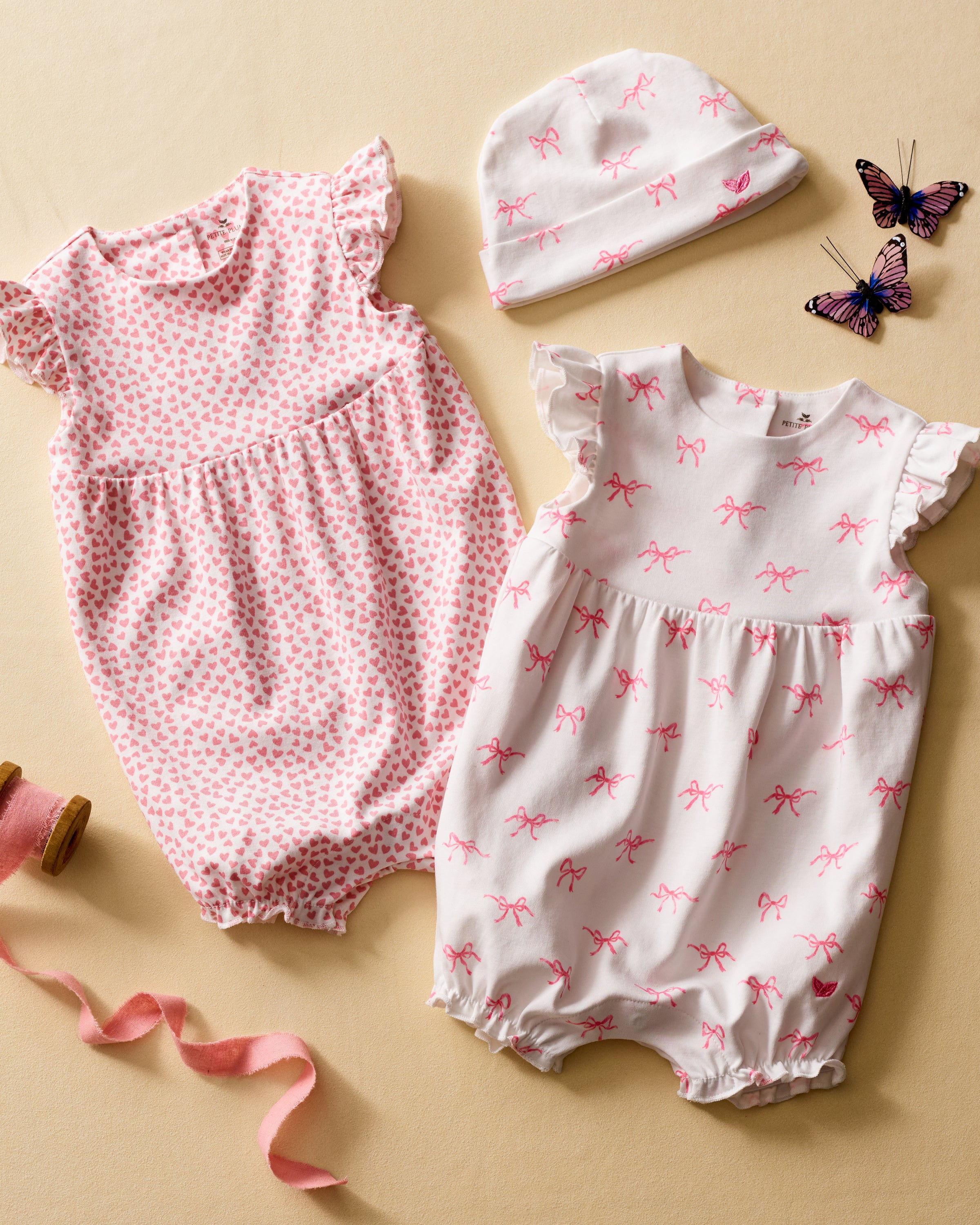 On a beige backdrop, two adorable Petite Plume rompers with ruffled sleeves are displayed. One romper has pink hearts, while the Babys Pima Isla Play Romper in Blushing Bows features pink bows. Crafted from Pima cotton, theyre paired with a matching hat, two purple butterflies, a pink ribbon, and pink thread.