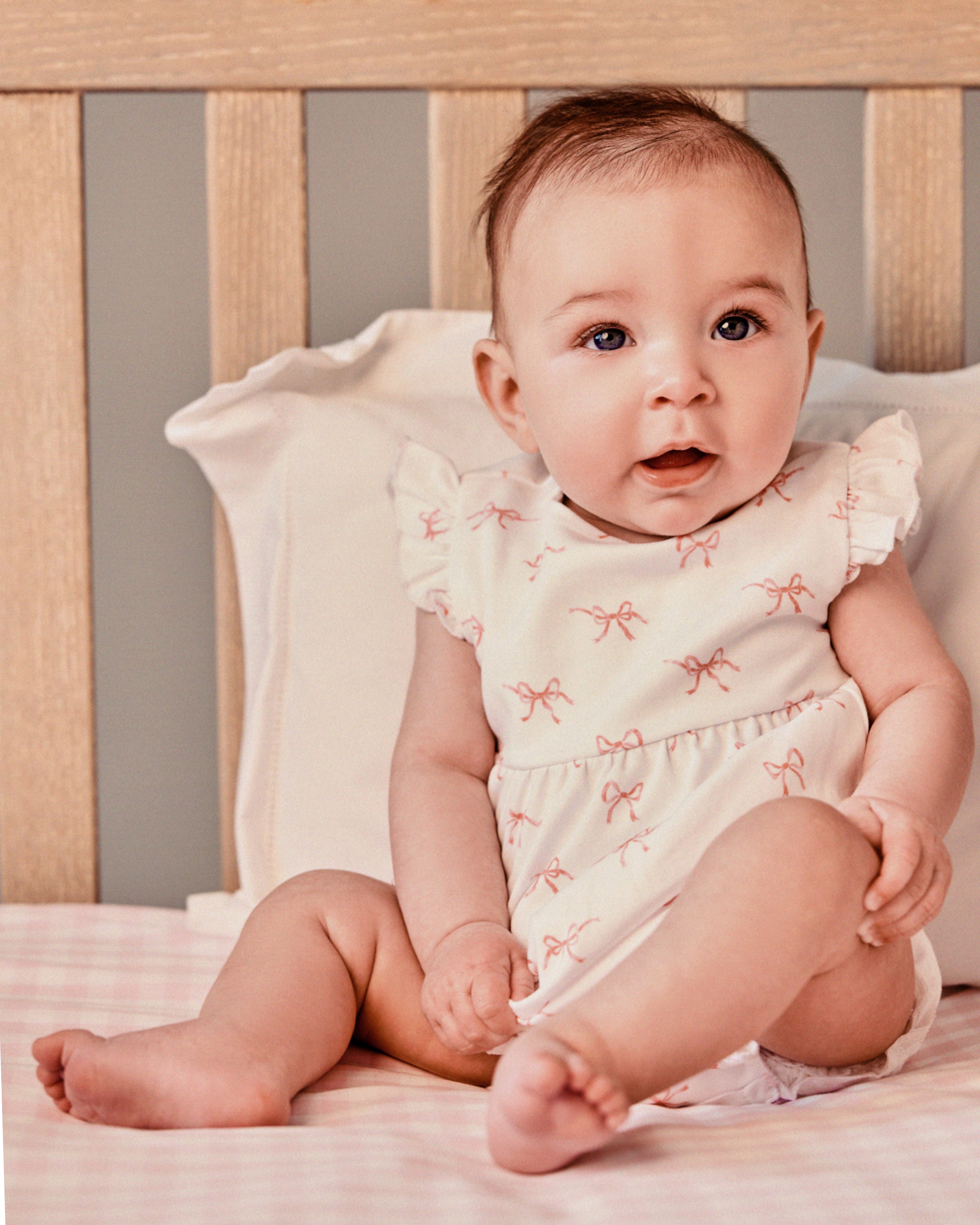 In a crib with wooden slats and pale pink bedding, a baby sits wearing the Petite Plume Babys Pima Isla Play Romper in Blushing Bows, its pink bow patterns enhancing her light brown hair and curious gaze.