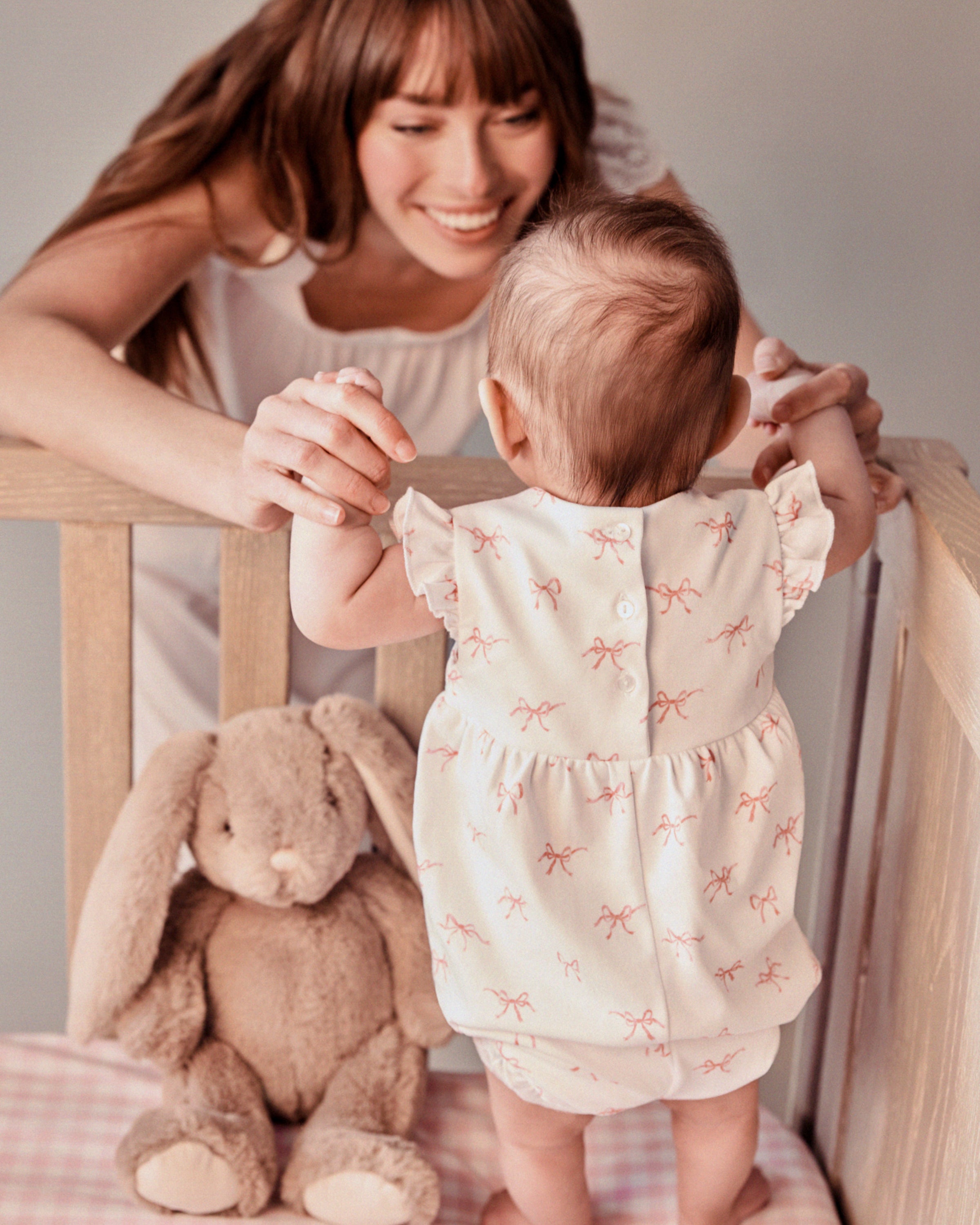 A woman smiles at a baby in a crib, wearing Petite Plumes Babys Pima Isla Play Romper in Blushing Bows. Nearby, a plush bunny sits among the pattern.