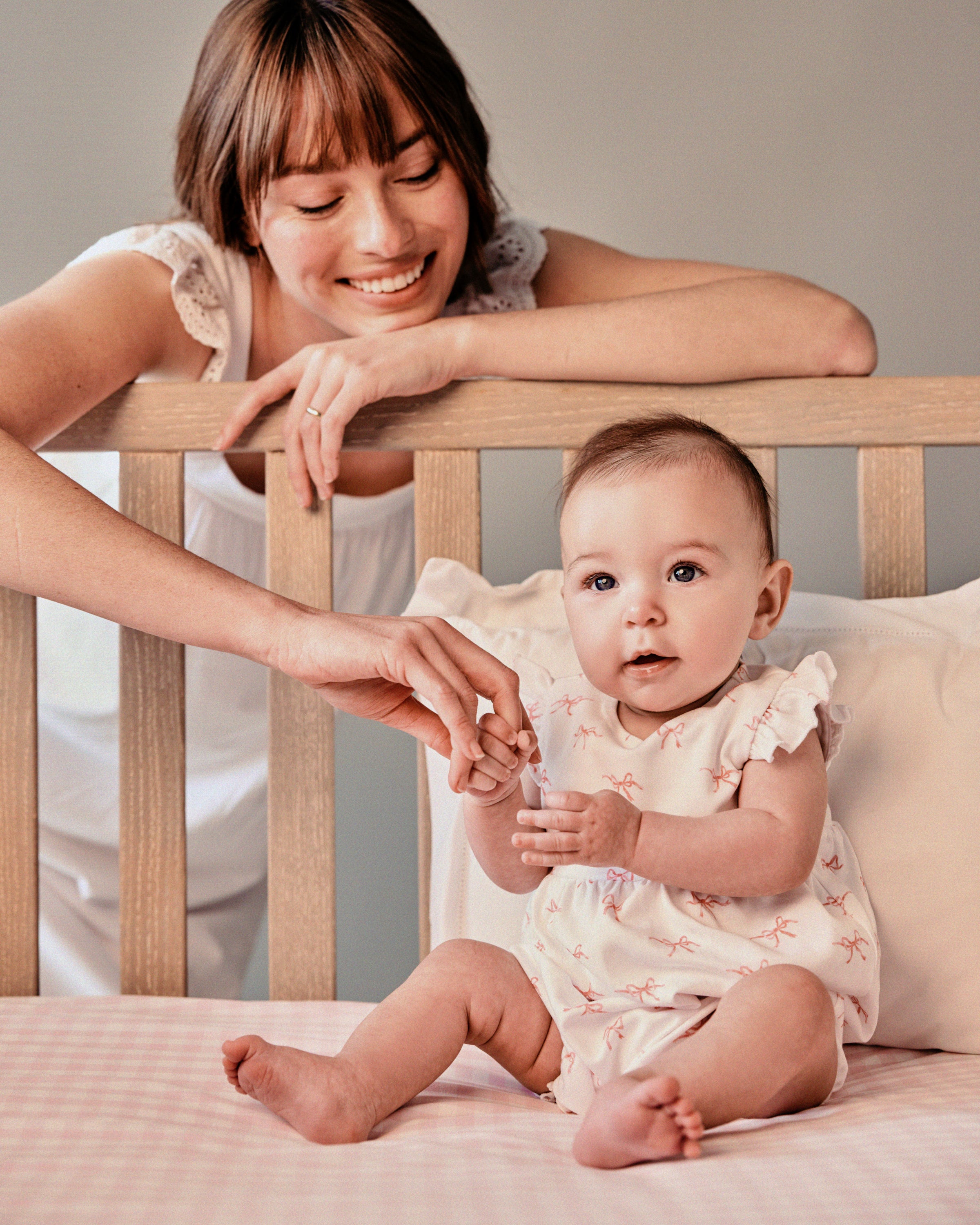 A woman smiles at a baby in a wooden crib on a pink checkered blanket. The baby, wearing Petite Plumes Babys Pima Isla Play Romper in Blushing Bows, gently touches the womans hand while looking at the camera.