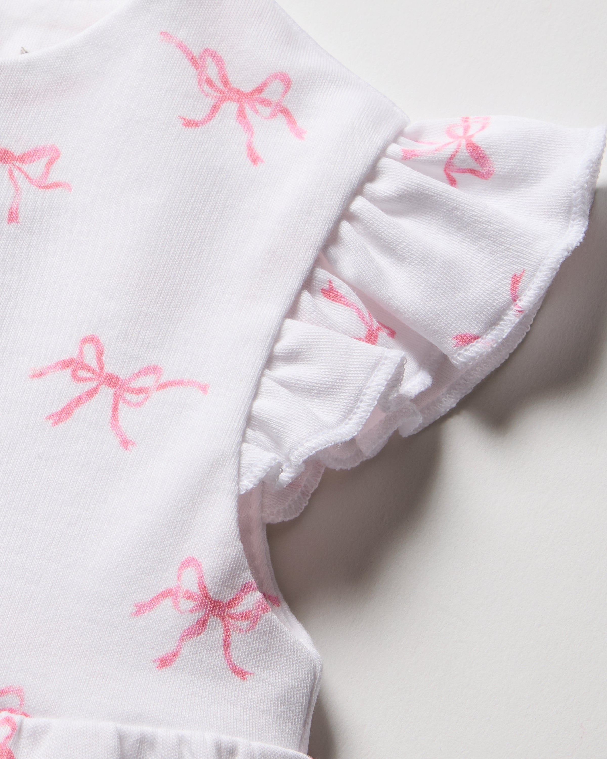 Close-up of Petite Plumes Babys Pima Isla Play Romper in Blushing Bows, featuring ruffled sleeves and crafted from soft Pima cotton. The white romper is adorned with blushing pink bow patterns, highlighting its delicate and feminine design.