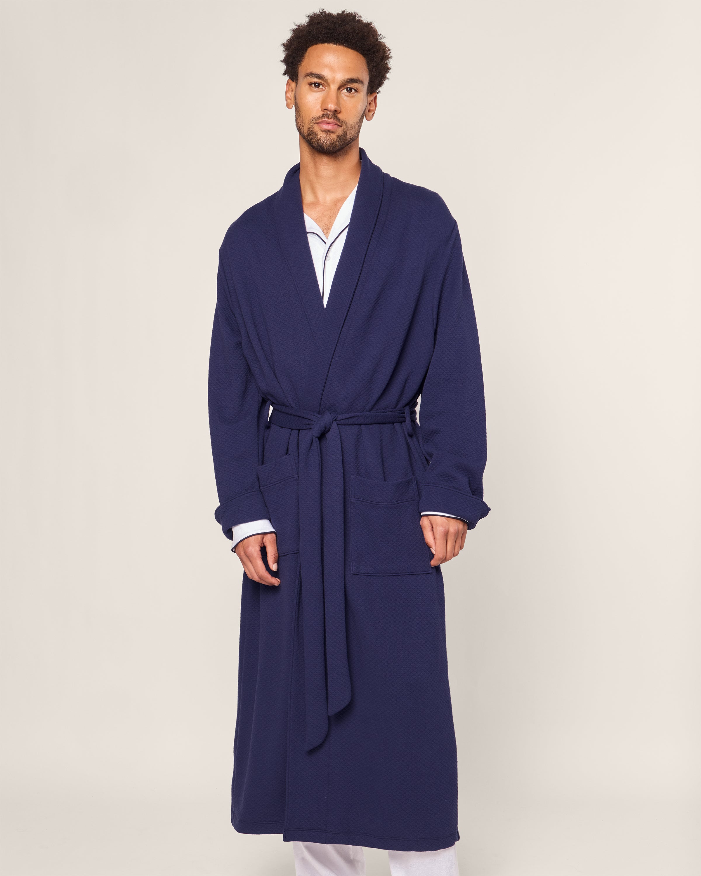 A man stands wearing the Petite Plume Mens Pima Robe in Blue Dusk Jacquard, featuring a quilted effect and waist belt. Underneath, he wears a white shirt and lighter pants. A plain beige background highlights the robes elegance.