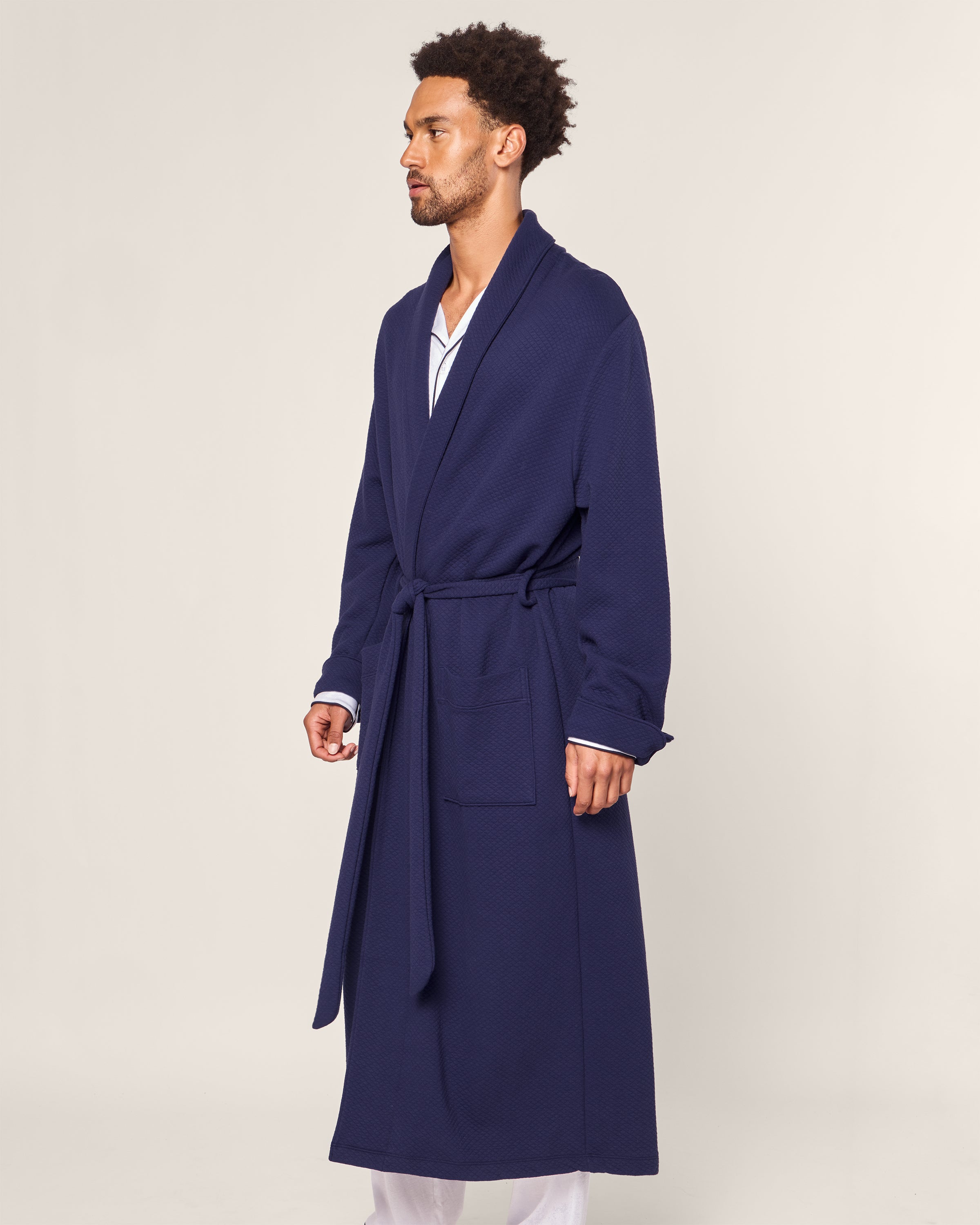 In a light gray setting, a man in profile dons the Petite Plume Mens Pima Robe in Blue Dusk Jacquard with a belt, showcasing its subtle quilted effect. Beneath, he pairs soft white pajama pants and a Pima cotton shirt.