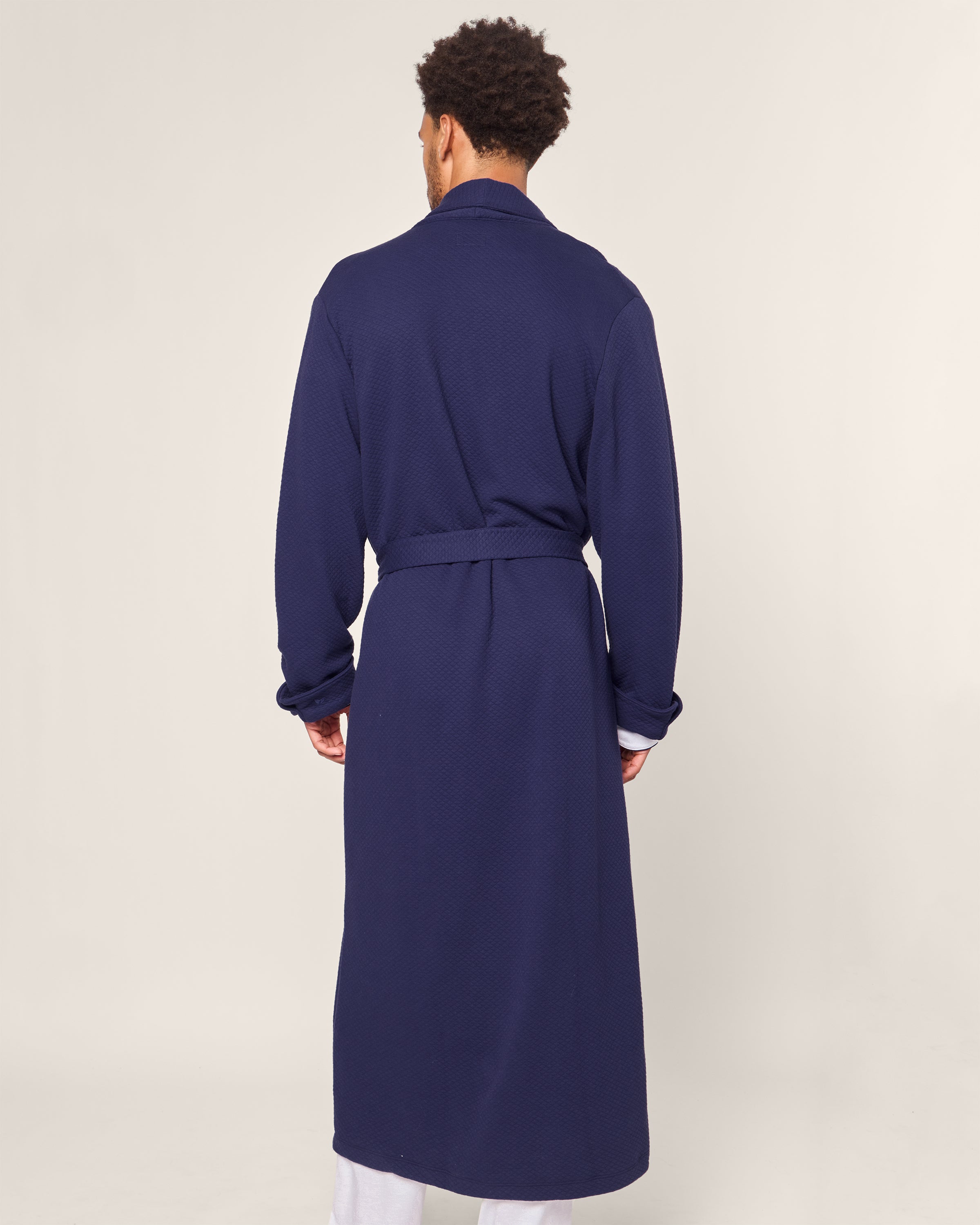 A person in a Mens Pima Robe in Blue Dusk Jacquard by Petite Plume, featuring a quilted effect belt tied at the waist, stands against a plain background, facing away from the camera.