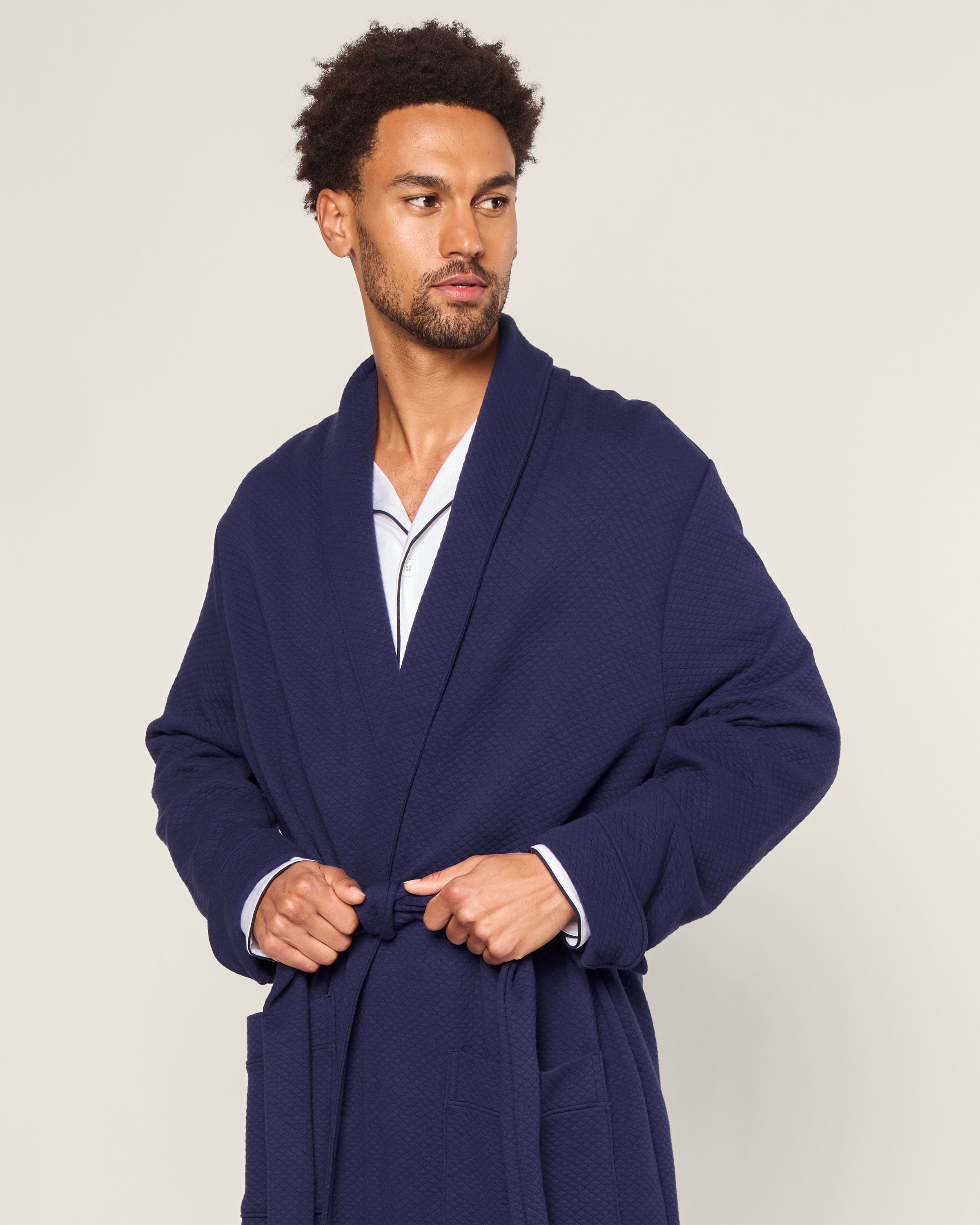 A man stands against a plain background, looking right, wearing Petite Plumes Mens Pima Robe in Blue Dusk Jacquard over light pajamas. He ties the belt at his waist, adding an elegant touch to his relaxed ensemble.