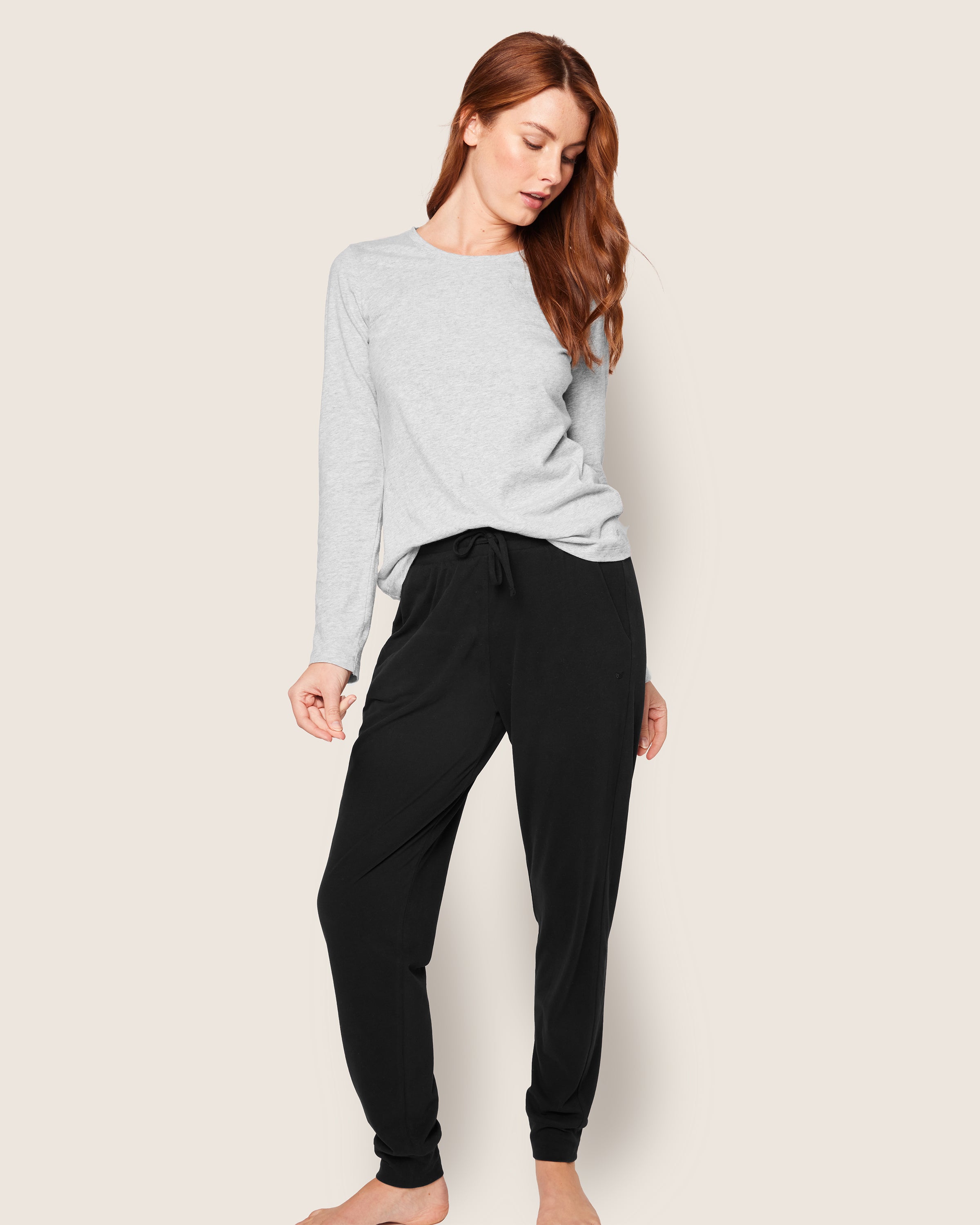 A woman with long red hair wears Petite Plumes Womens Pima Lounge Pants in Black, made from luxurious Peruvian Pima cotton. She stands against a plain background, looking down with a relaxed pose, her arms slightly bent.