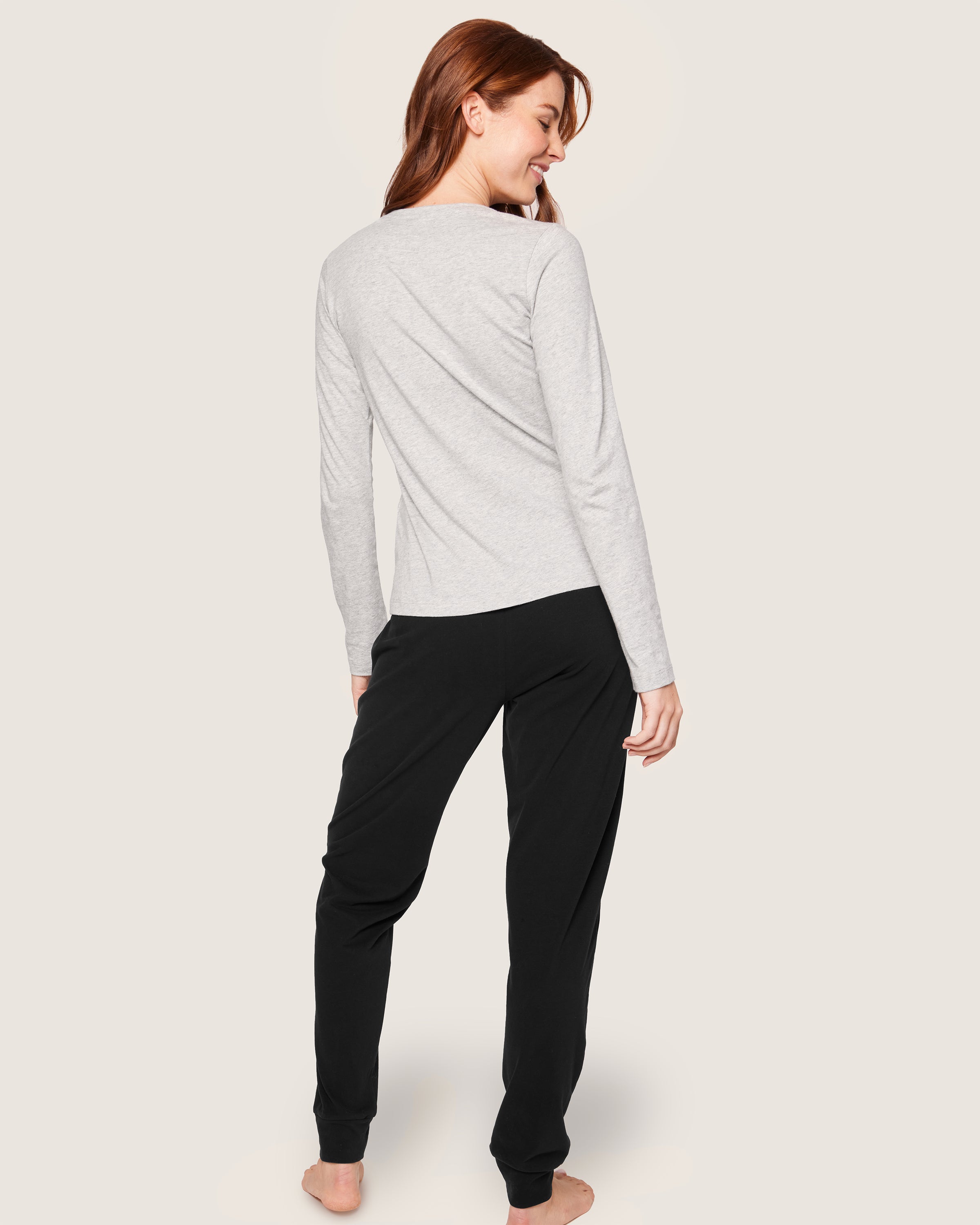 A woman with long brown hair stands barefoot, facing away, wearing a light gray, long-sleeve shirt made from luxurious Peruvian Pima cotton paired with Petite Plumes Womens Pima Lounge Pants in Black. The plain beige background enhances the elegance of her attire.