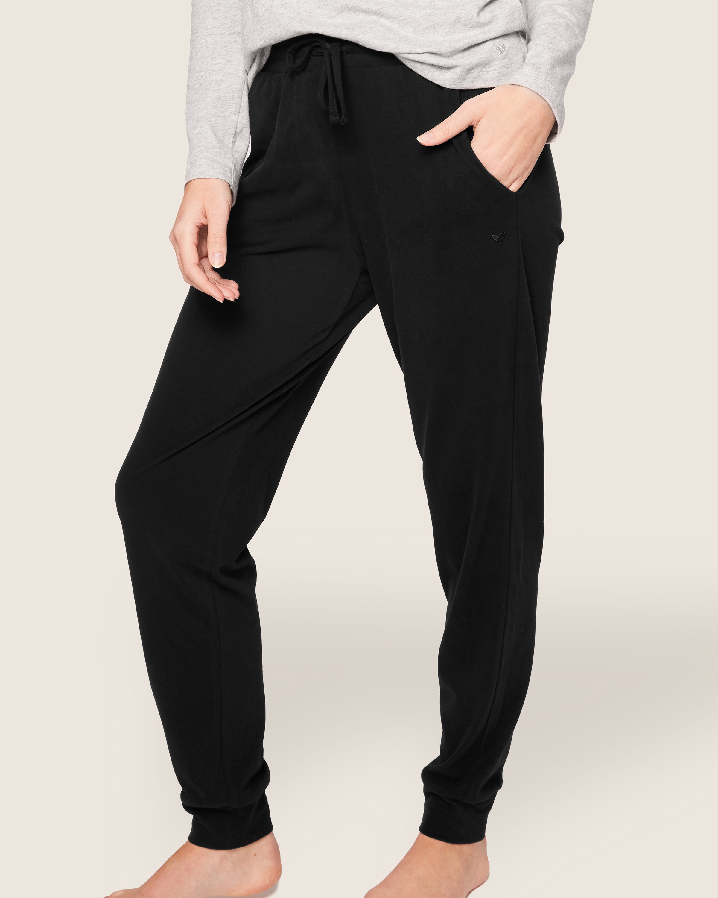 A person wearing Petite Plumes Womens Pima Lounge Pants in Black has hands partially in pockets. They sport a light gray long-sleeve top of luxurious Peruvian Pima cotton and are barefoot against a plain off-white background.