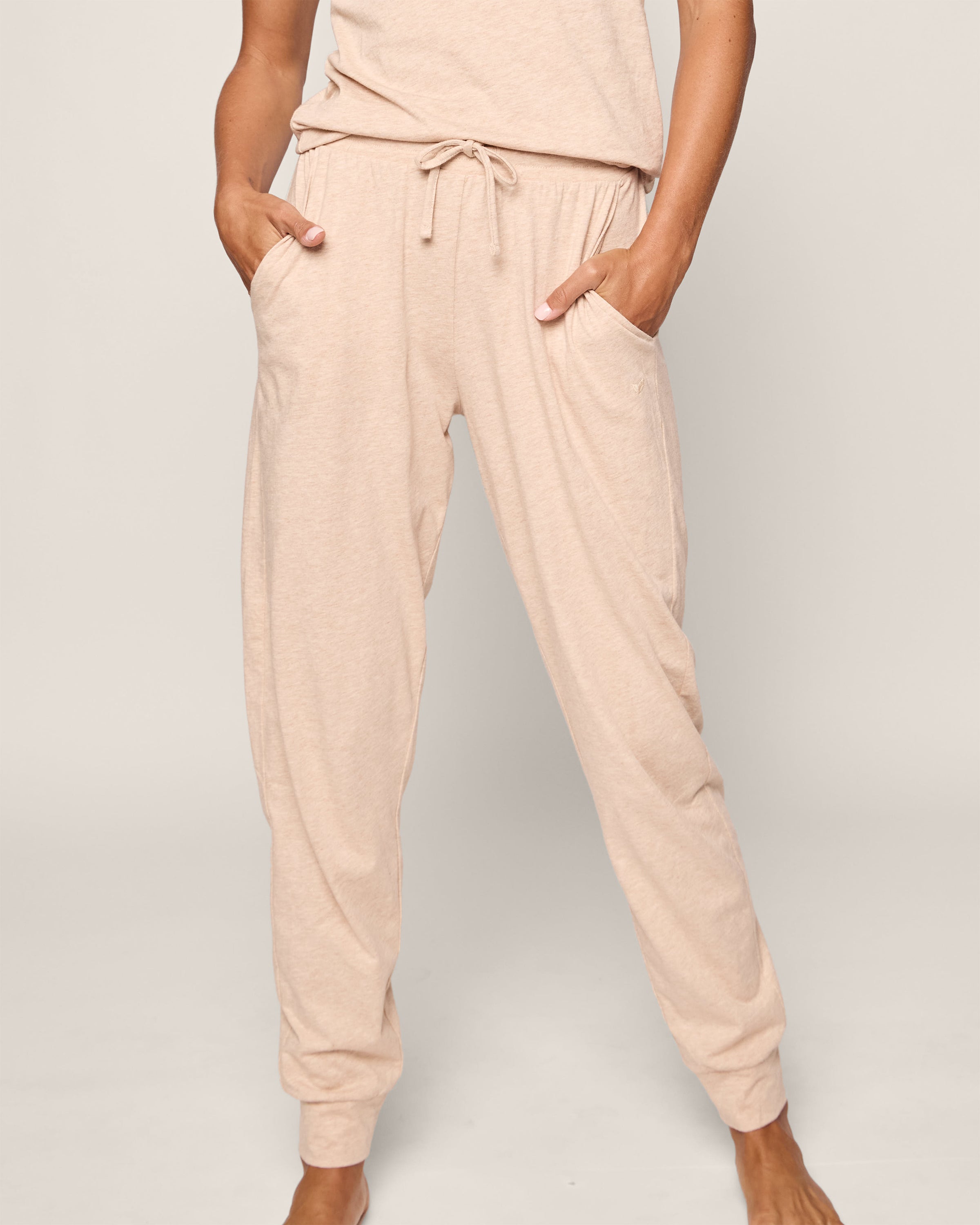 A person is wearing Petite Plumes Womens Pima Lounge Pants in Camel, made of luxurious Peruvian Pima cotton with a drawstring waist and hands in pockets. Their upper body and head are not visible against a plain, light background.