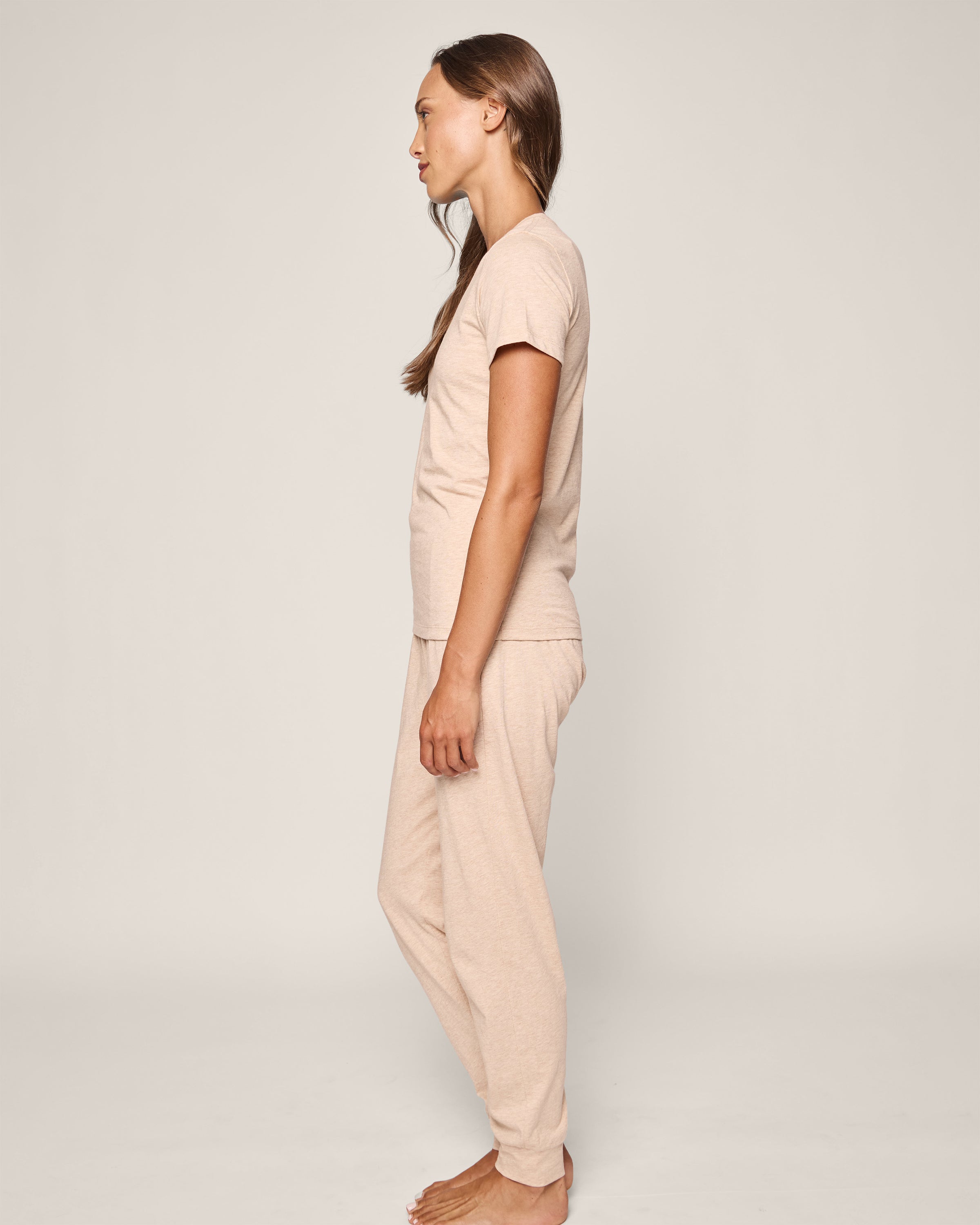 A woman stands sideways, facing left in Petite Plumes Womens Pima Lounge Pants in camel and a matching beige T-shirt. She is barefoot on a plain white background, with straight hair cascading down her back.