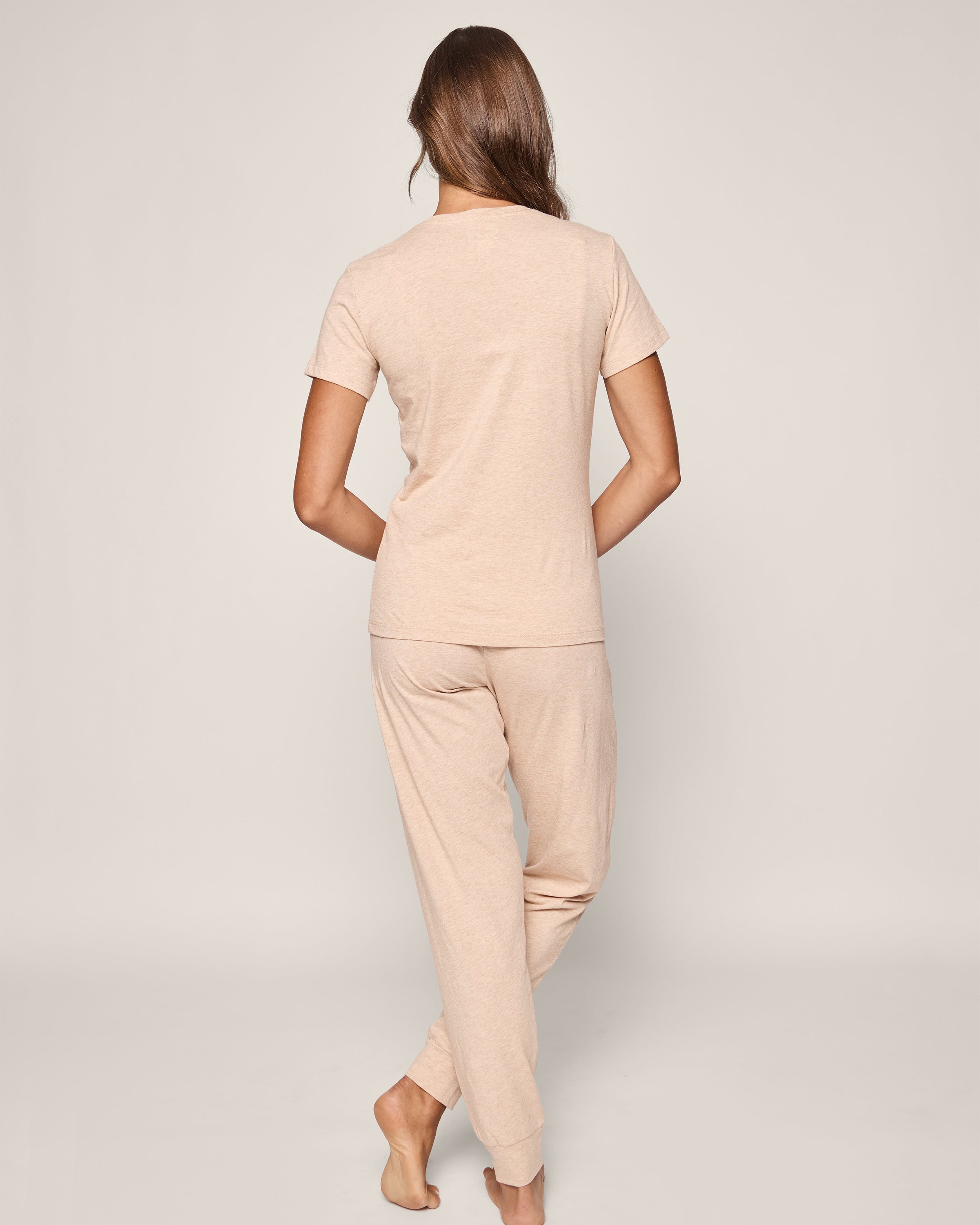 A woman with long hair, seen from behind, is barefoot on a light background. Shes wearing Womens Pima Lounge Pants in Camel by Petite Plume, paired with a matching short-sleeve top made of luxurious Peruvian Pima cotton, her hands clasped behind her back.