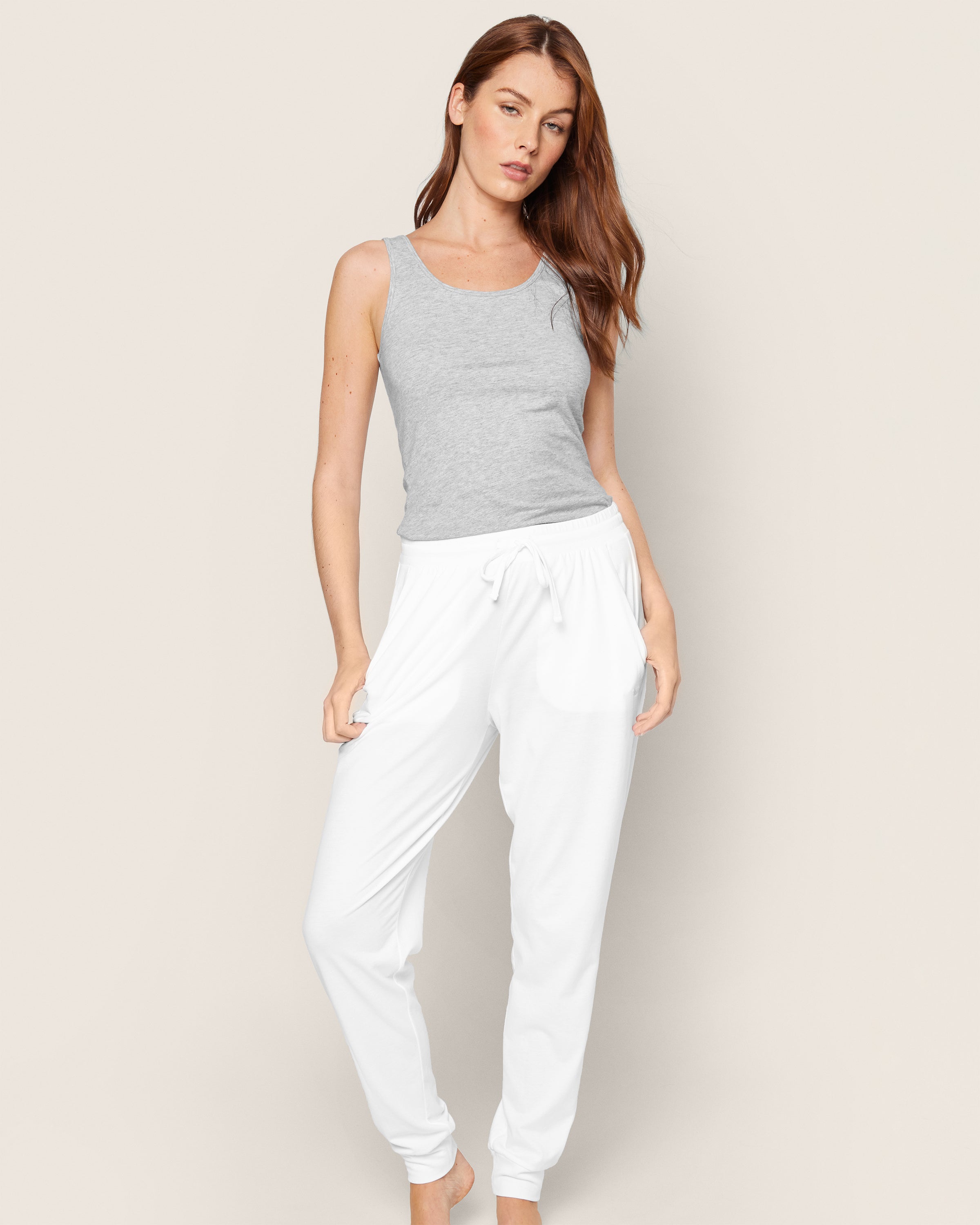 A person with long hair wears Petite Plumes Womens Pima Lounge Pants in white and a gray tank top, against a light beige background. They lean slightly in a relaxed pose, embodying casual elegance and the simplicity of bedtime rituals.