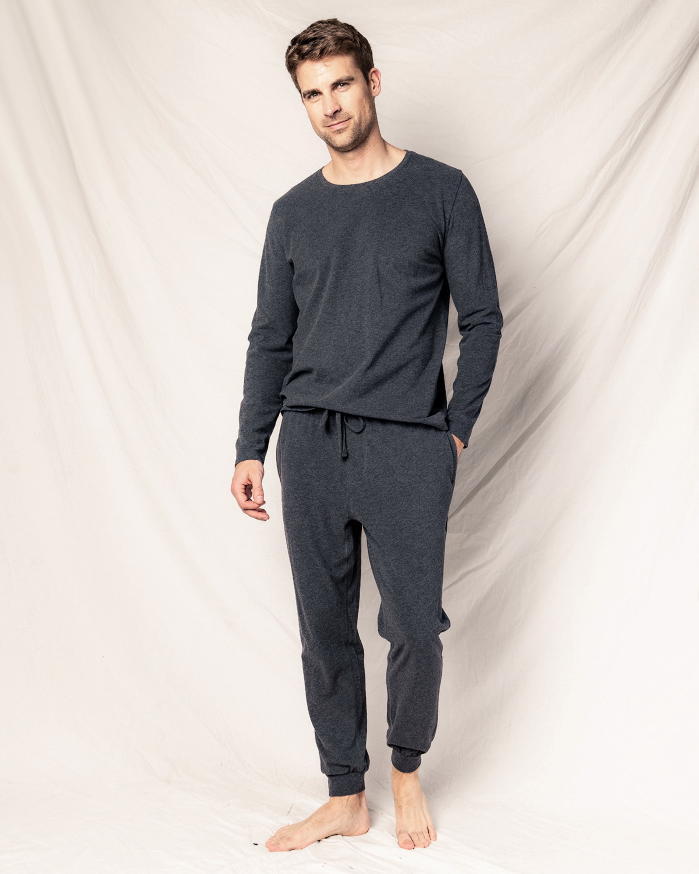 Barefoot against a light backdrop, a man wears Petite Plumes Mens Pima Pants in Dark Heather Grey, made from luxurious jersey knit Peruvian Pima cotton. The loungewear set includes long sleeves and jogger pants; he has short hair, a slight smile, and one hand in his pocket.