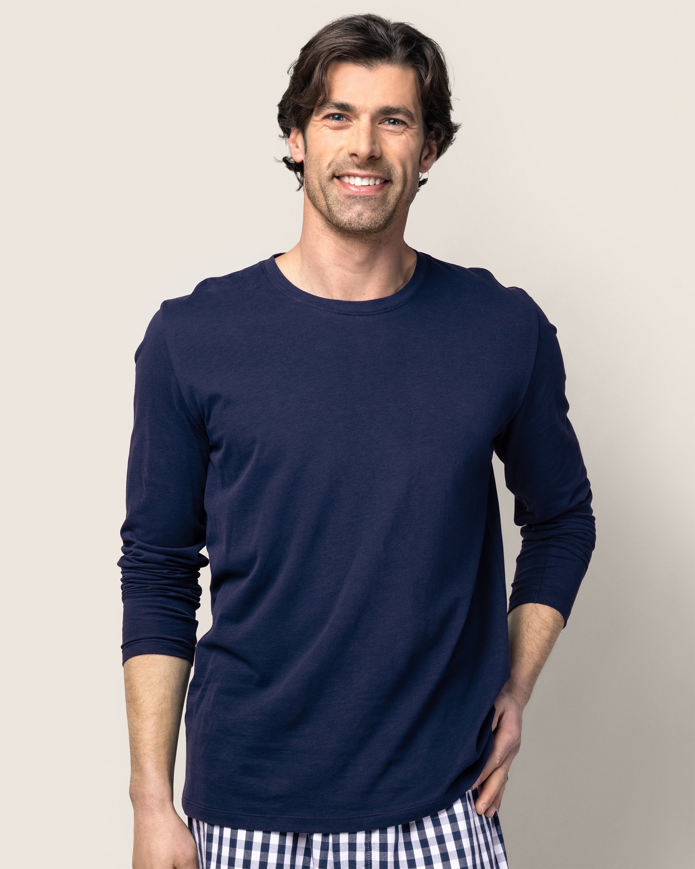 A man with shoulder-length brown hair and a beard smiles while wearing the navy Men’s Pima Long Sleeve T-shirt Duo by Petite Plume and plaid pajama pants of soft Peruvian Pima cotton. Standing against a neutral background, hand in pocket, he embodies his relaxing bedtime ritual.