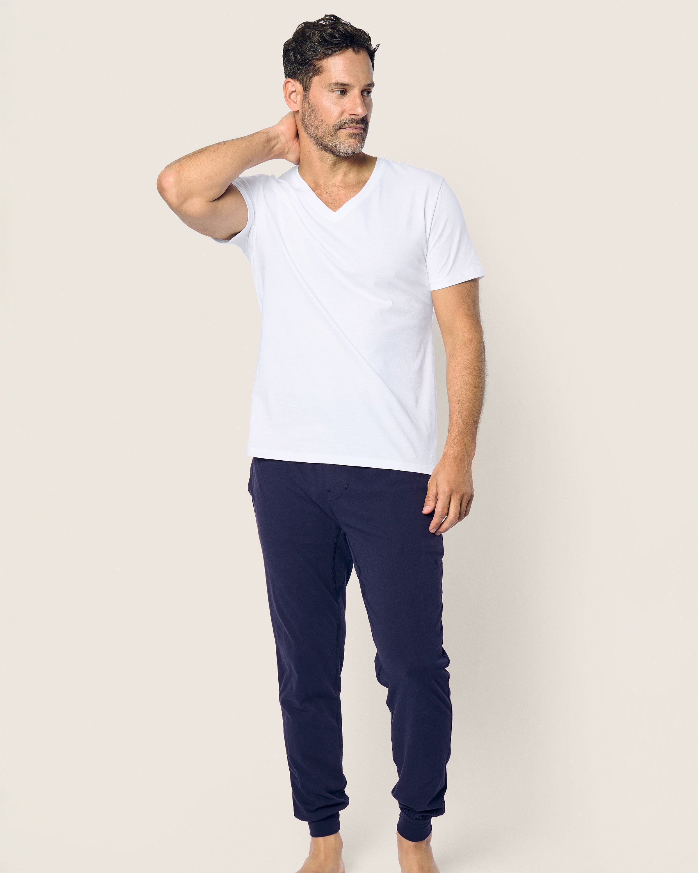 Against a light background, a man with short dark hair wearing a white T-shirt and Mens Pima Pants in Navy by Petite Plume, made from luxurious jersey knit fabric, stands barefoot, looking to the side and touching the back of his neck with one hand.