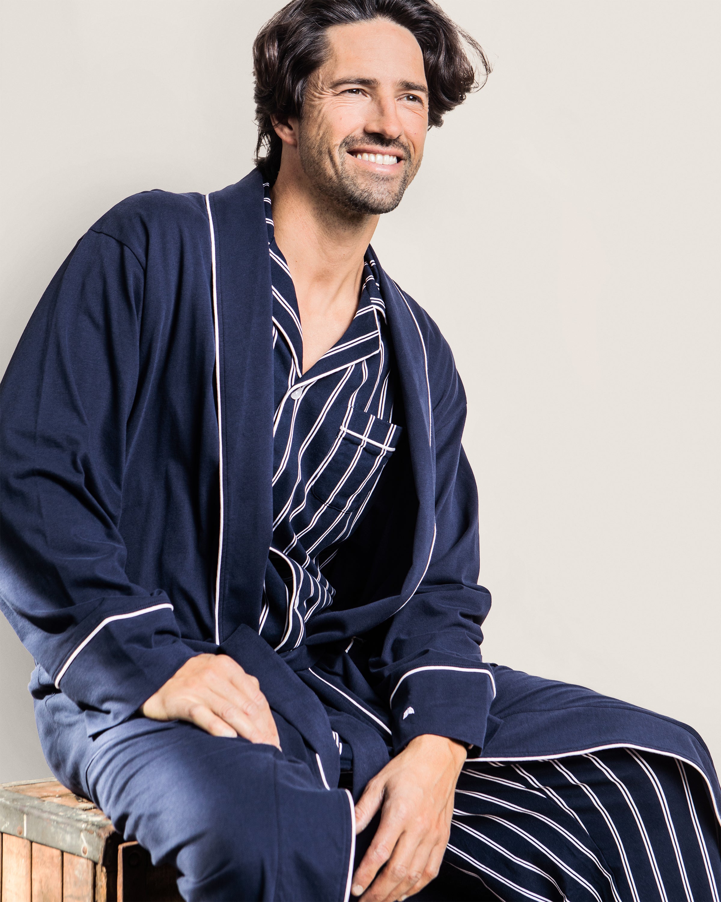 A smiling man wearing the Petite Plume Mens Pima Robe in Navy with white trim sits on a wooden block against a plain background.