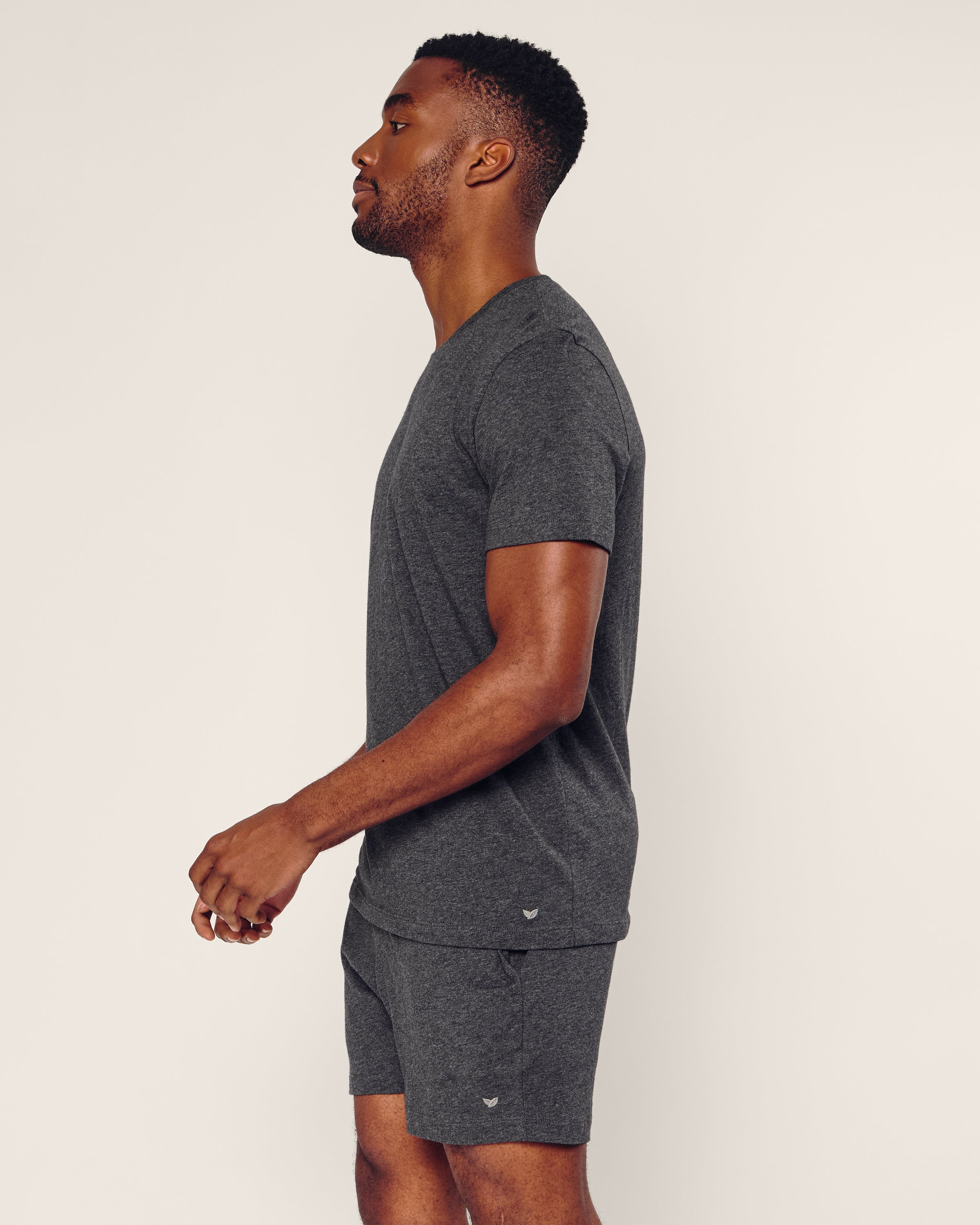 A man poses in profile against a plain background, wearing Petite Plumes dark grey Pima shorts. The set includes a short-sleeve shirt and matching shorts, crafted from soft Peruvian Pima cotton.