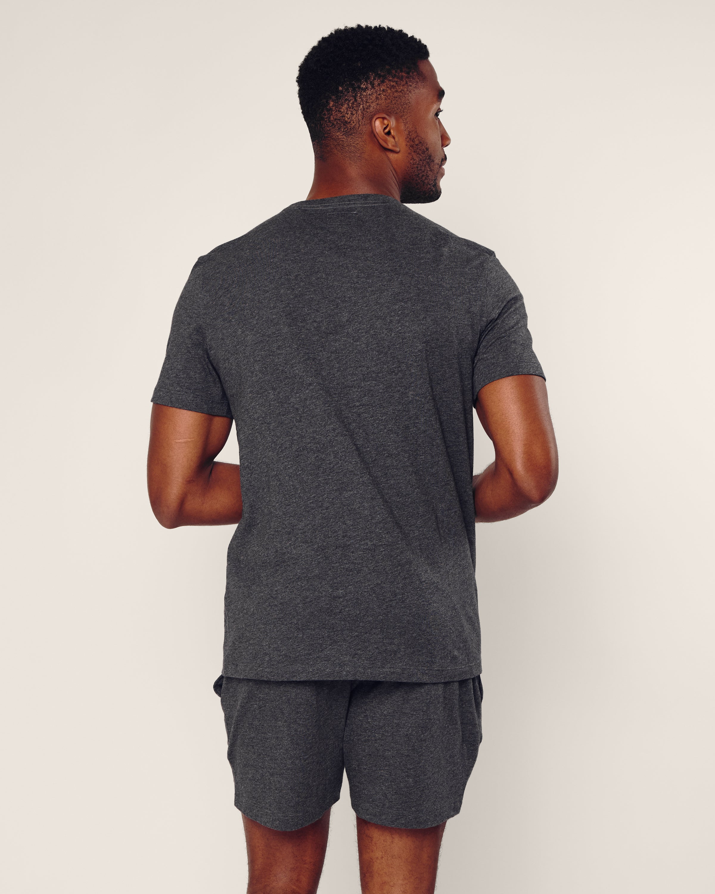 A man stands with his back to the camera, wearing Petite Plumes Mens 5 Pima Shorts in Dark Grey. The soft Peruvian Pima cotton shorts complement a matching dark gray t-shirt against a plain, light backdrop, emphasizing their casual fit and texture.