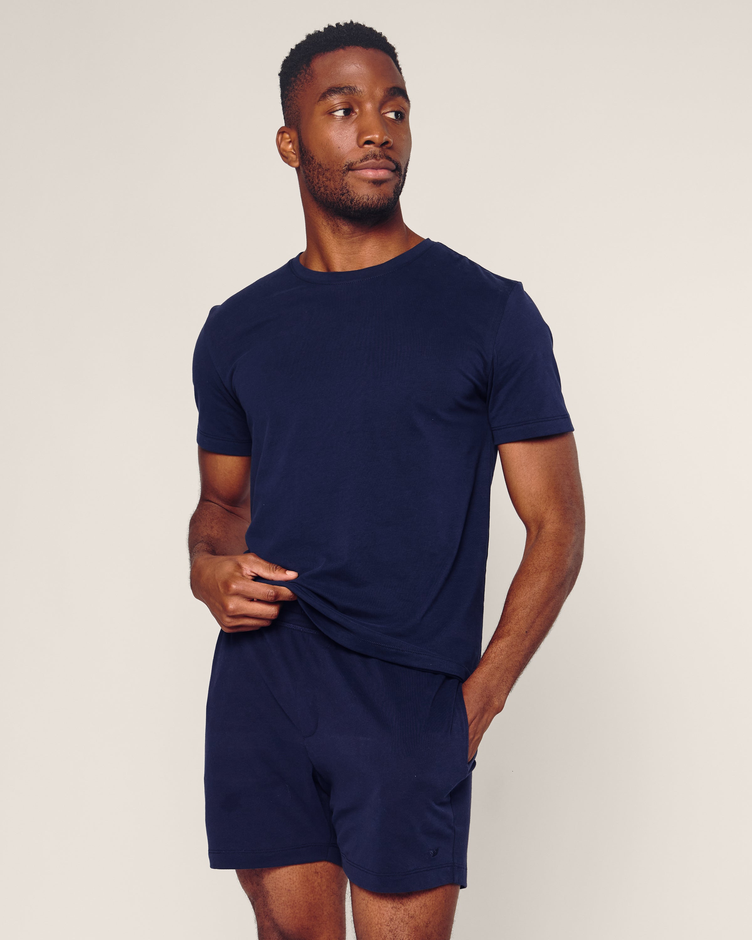 A person with a short beard stands against a plain background, wearing Petite Plumes Mens 5 Pima Shorts in Navy with a matching Peruvian Pima cotton navy blue t-shirt. They look relaxed, gazing slightly to the side, one hand holding the shirt hem and the other in the shorts pocket.