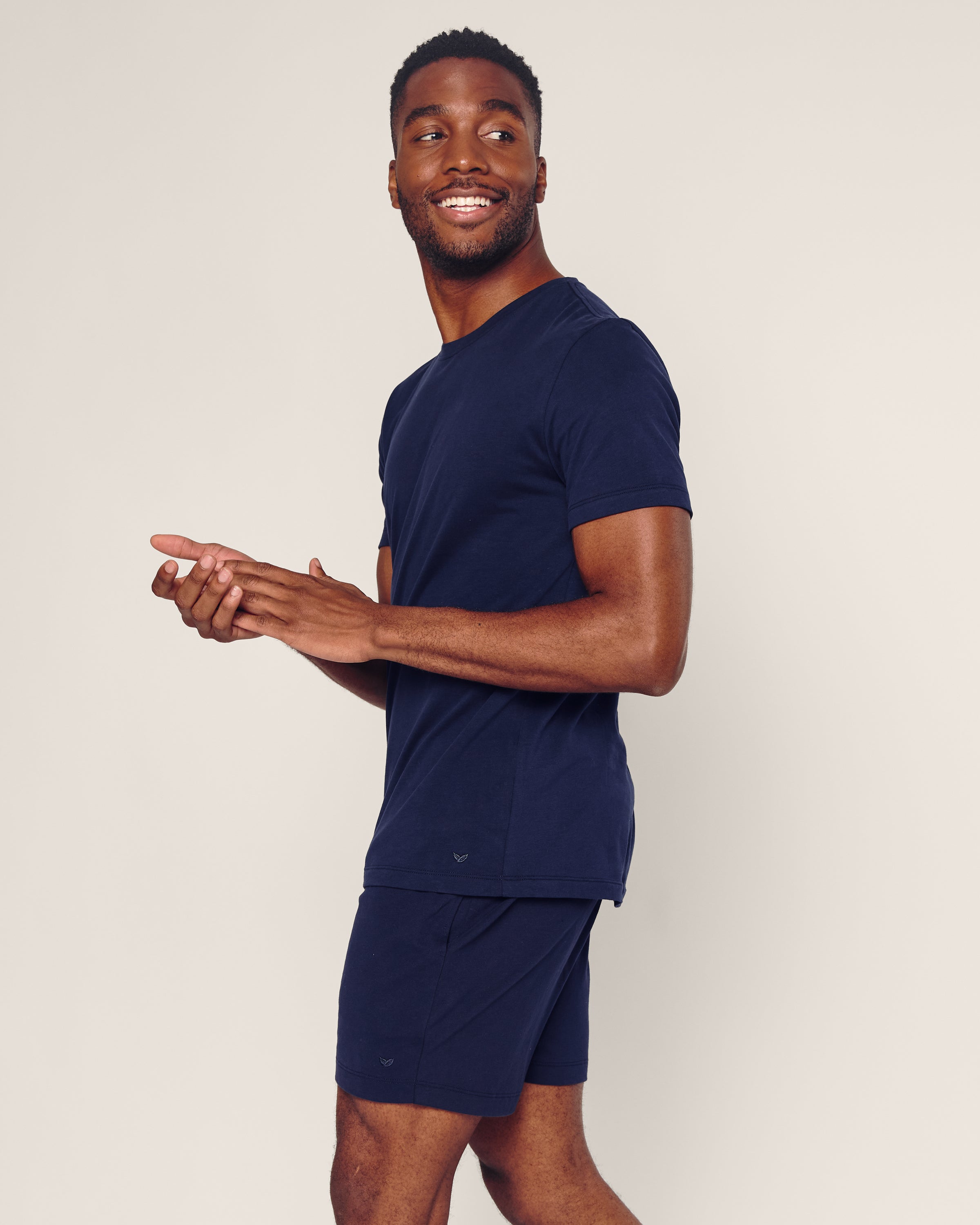 A man smiles, looking right while wearing Petite Plumes Mens 5 Pima Shorts in Navy and a matching t-shirt, made from soft Peruvian Pima cotton. He stands against a light-colored background with his hands clasped in front.