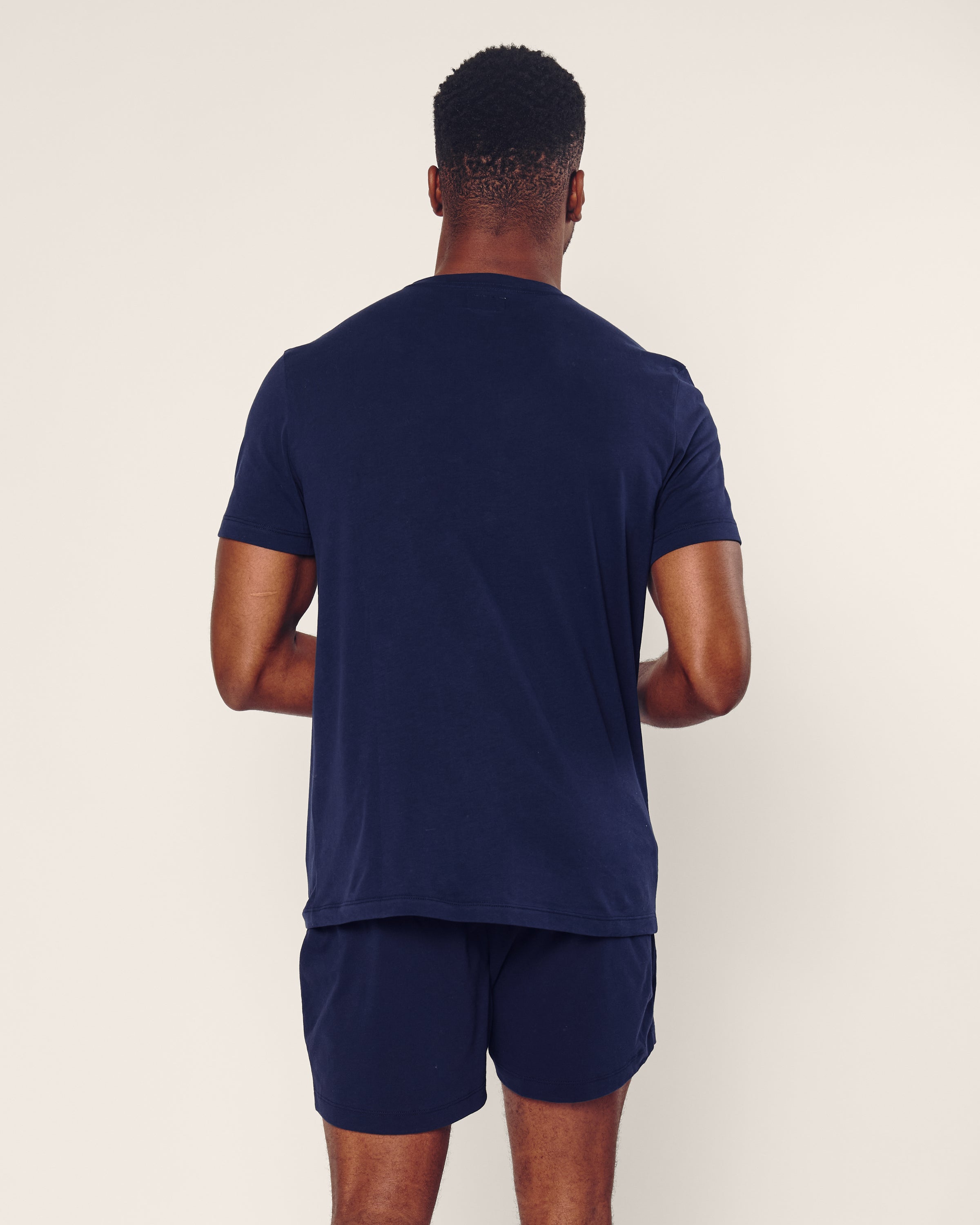 A person with short hair stands facing away, wearing Petite Plumes Mens 5 Pima Shorts in Navy and a matching navy blue T-shirt made of soft jersey knit fabric against a plain light background.