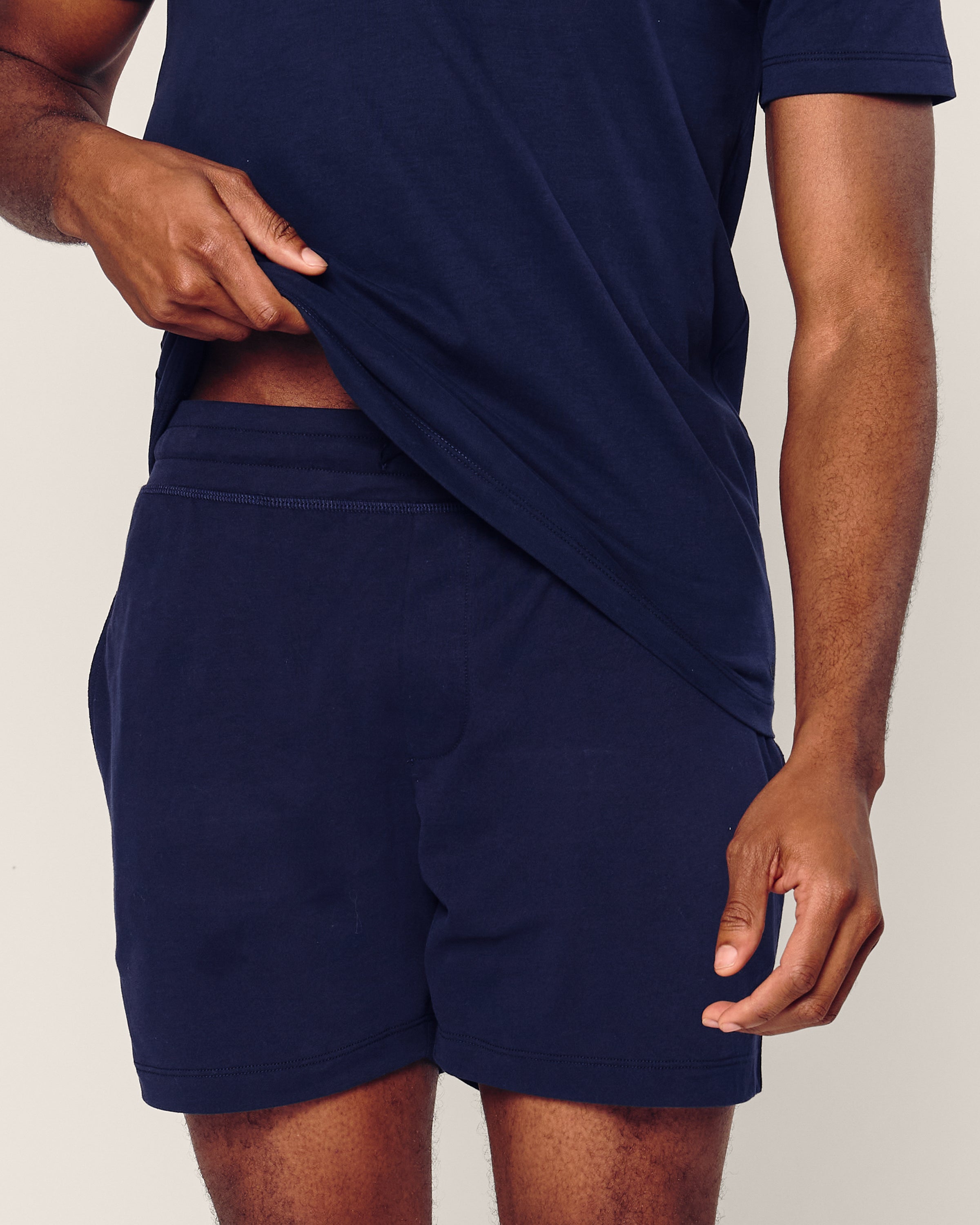 A person models the Mens 5 Pima Shorts by Petite Plume, slightly lifting their navy jersey knit T-shirt to reveal the shorts waistband while keeping one hand relaxed by their side against a neutral backdrop.