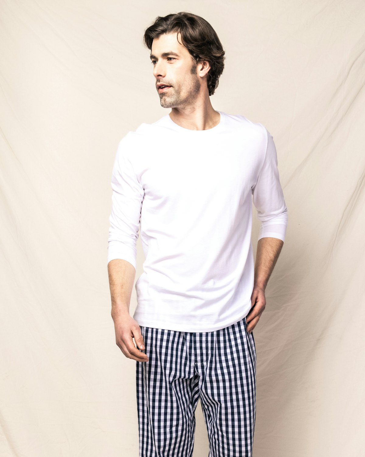 A man with medium-length hair wears the Men’s Pima Long Sleeve T-shirt Duo by Petite Plume in white, paired with black-and-white checkered pants. He stands against a beige backdrop, looking to the side with a relaxed posture.