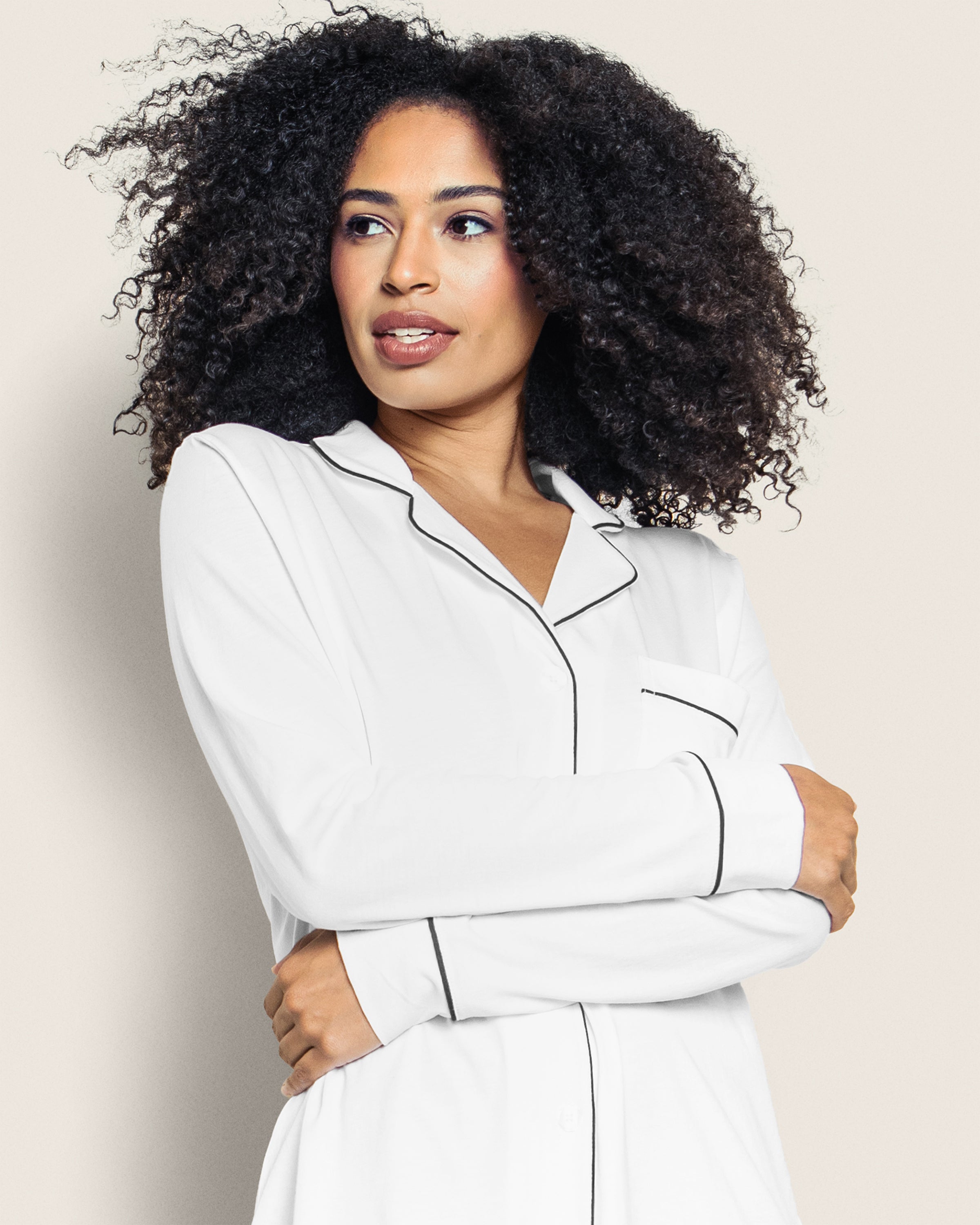 A woman with curly hair models the Womens Pima Nightshirt in White with Black Piping by Petite Plume, standing against a light background, looking to the side with her arms crossed.