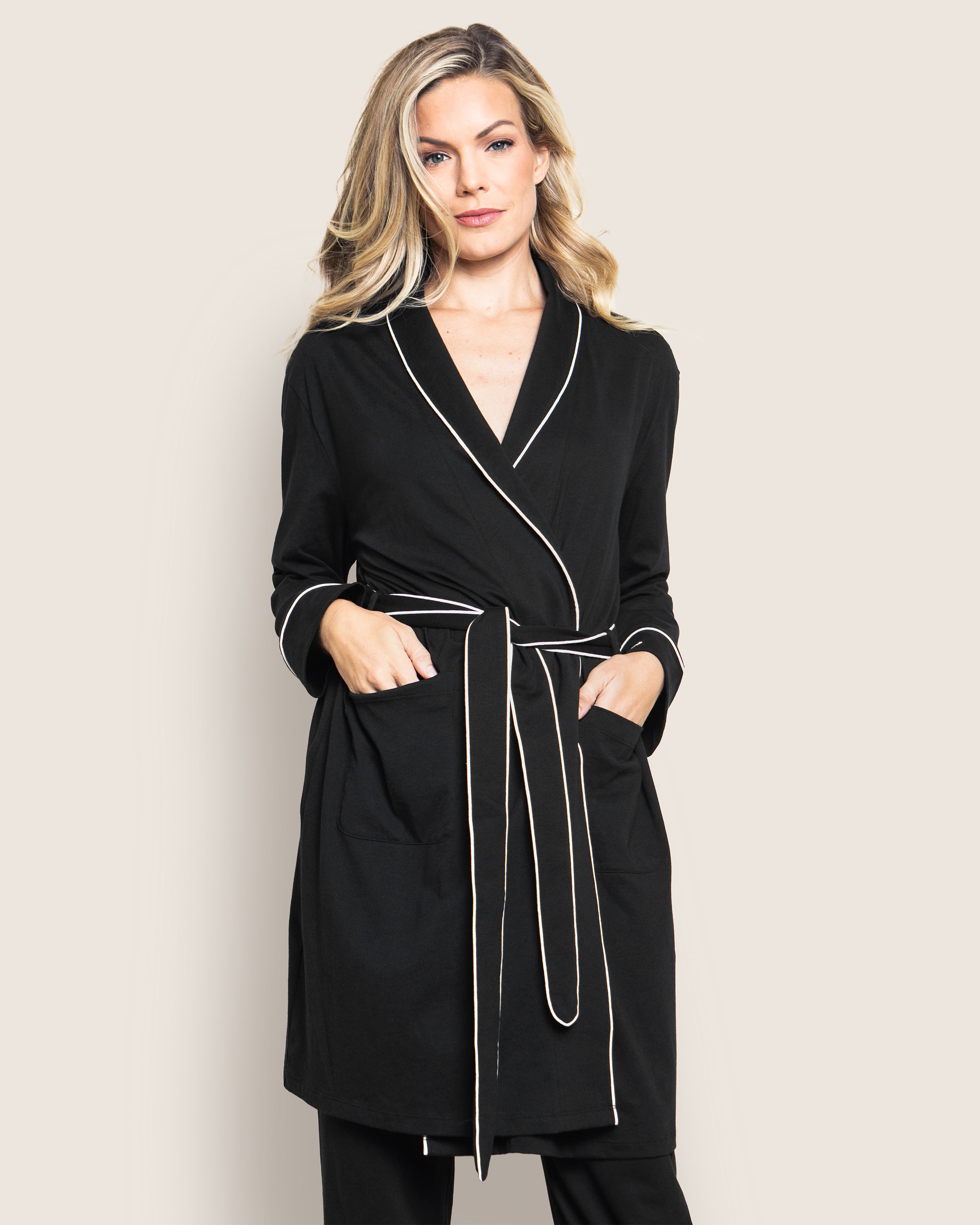 A woman wears a Petite Plume Womens Pima Robe in Black, characterized by white piping, long sleeves, and a waist tie. Made from soft Peruvian Pima cotton, she rests her hands in the front pockets while standing against a plain light gray background.