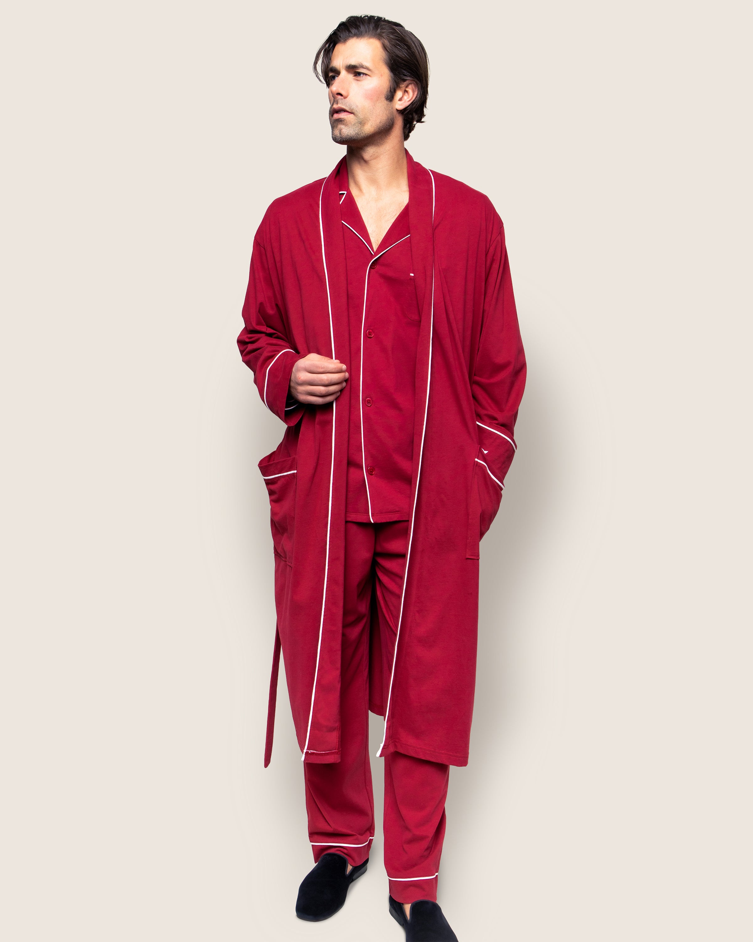 A man in a red Petite Plume Mens Pima Robe in Bordeaux, featuring classic silhouettes and white piping, stands against a plain background, hands slipping into his robe pockets. He gazes to the side, comfortably adorned in black slippers.